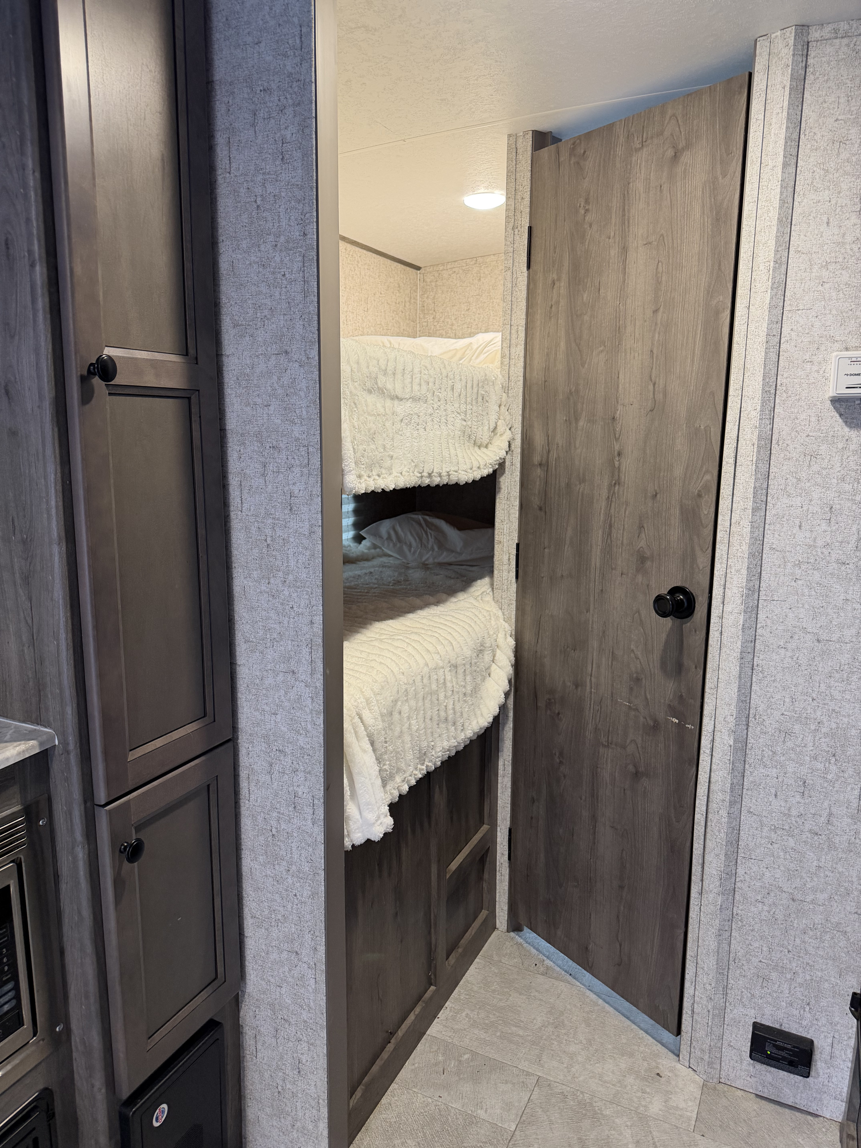 RV rental interior: A peek at cozy bunk beds with white bedding through a doorway, flanked by wood-look storage and a closed door.. Coachmen Apex Nano 2022