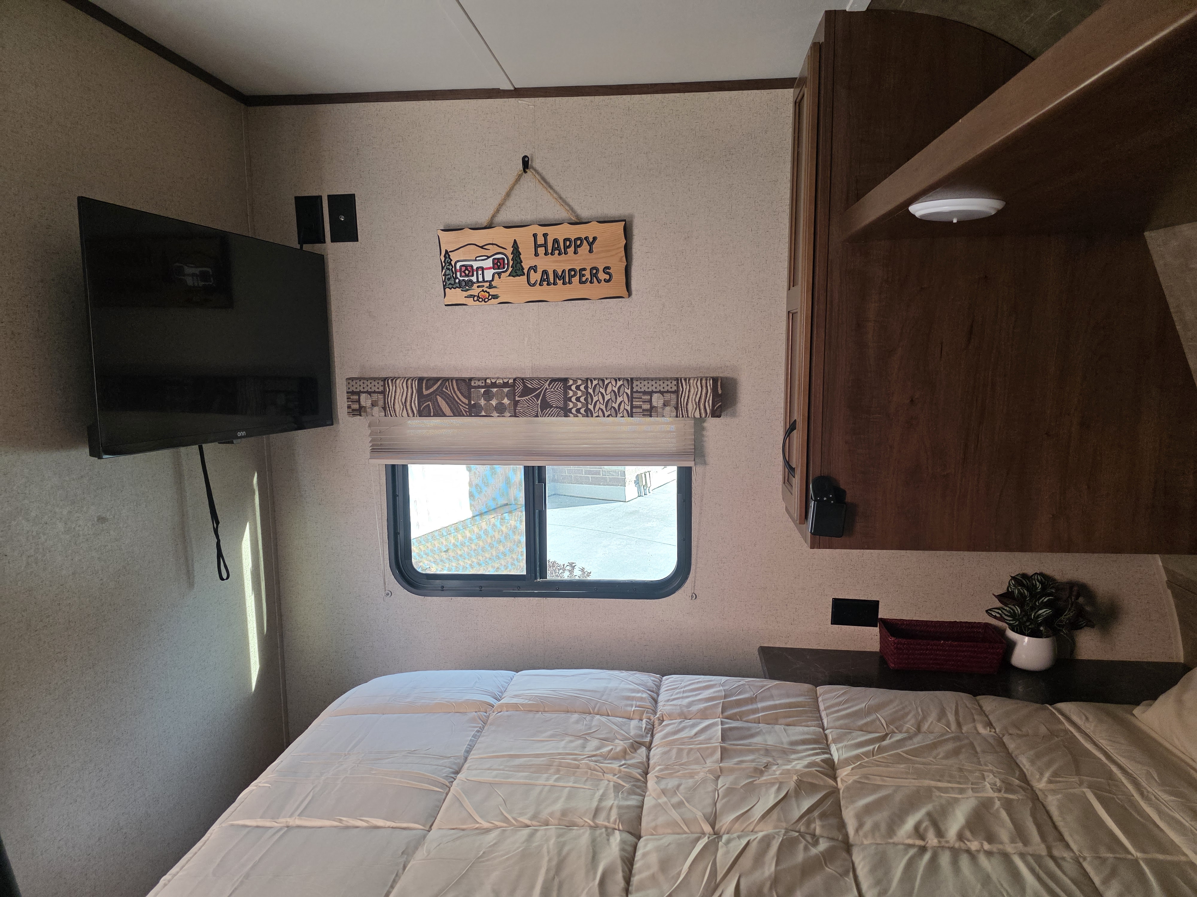 Cozy RV rental bedroom featuring a mounted TV, window with blinds, "Happy Campers" sign, and wood cabinets.. Jayco Jay Flight SLX 2016