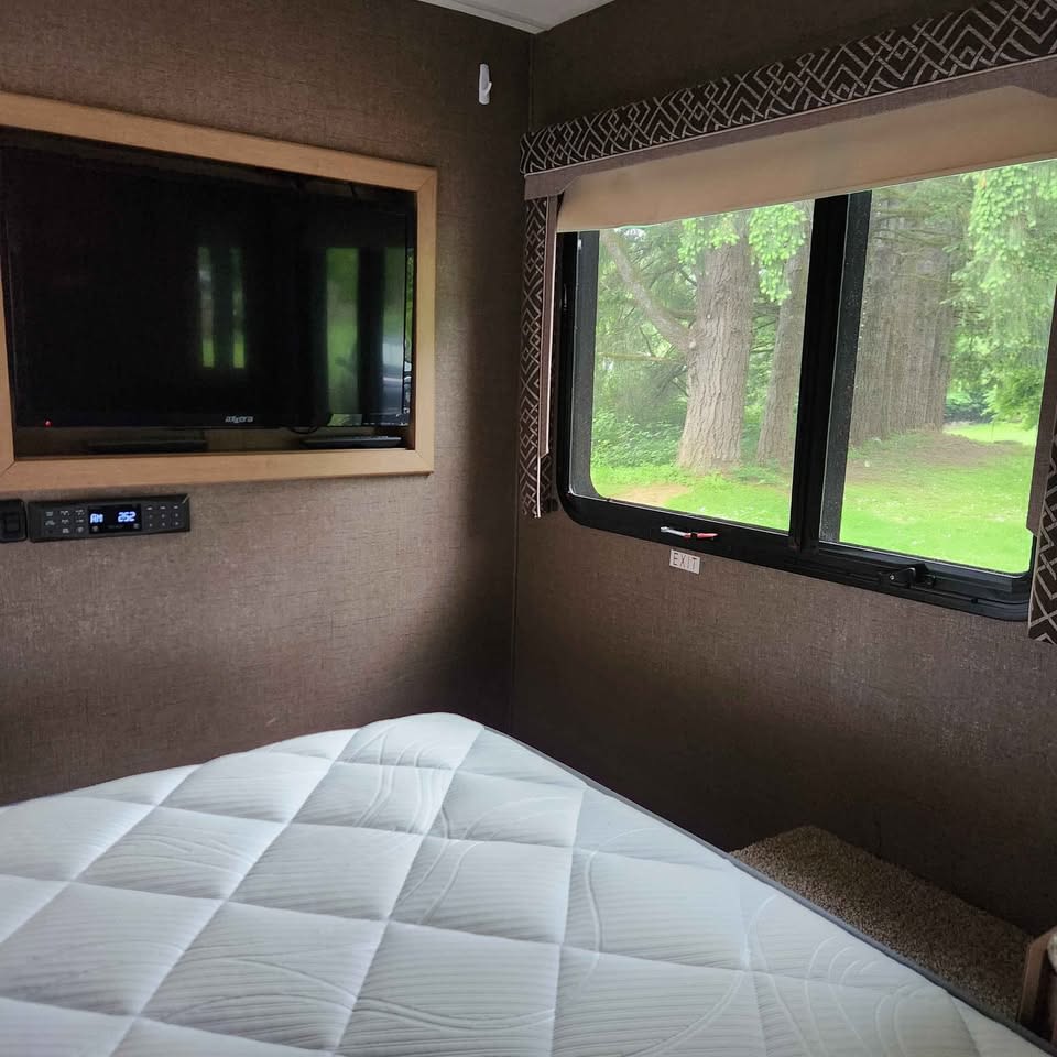 Cozy RV rental bedroom features a comfy quilted mattress, wall-mounted TV, and window framing lush green trees. Audio panel visible.. Thor Four Winds 2018