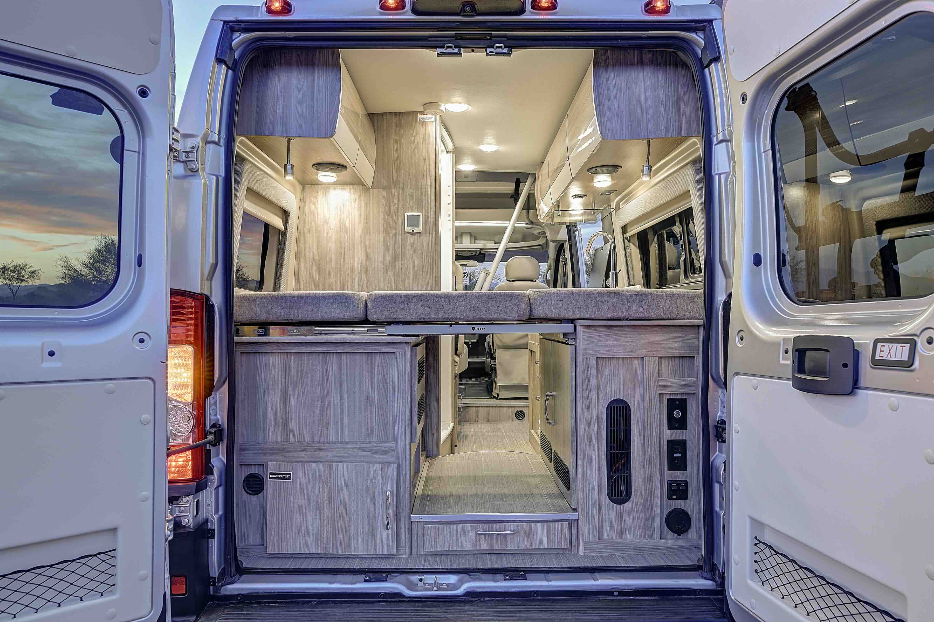 Inside a modern rv rental camper van: comfortable bed, kitchen area, and ample storage, viewed through open rear doors.. Thor Scope 2023
