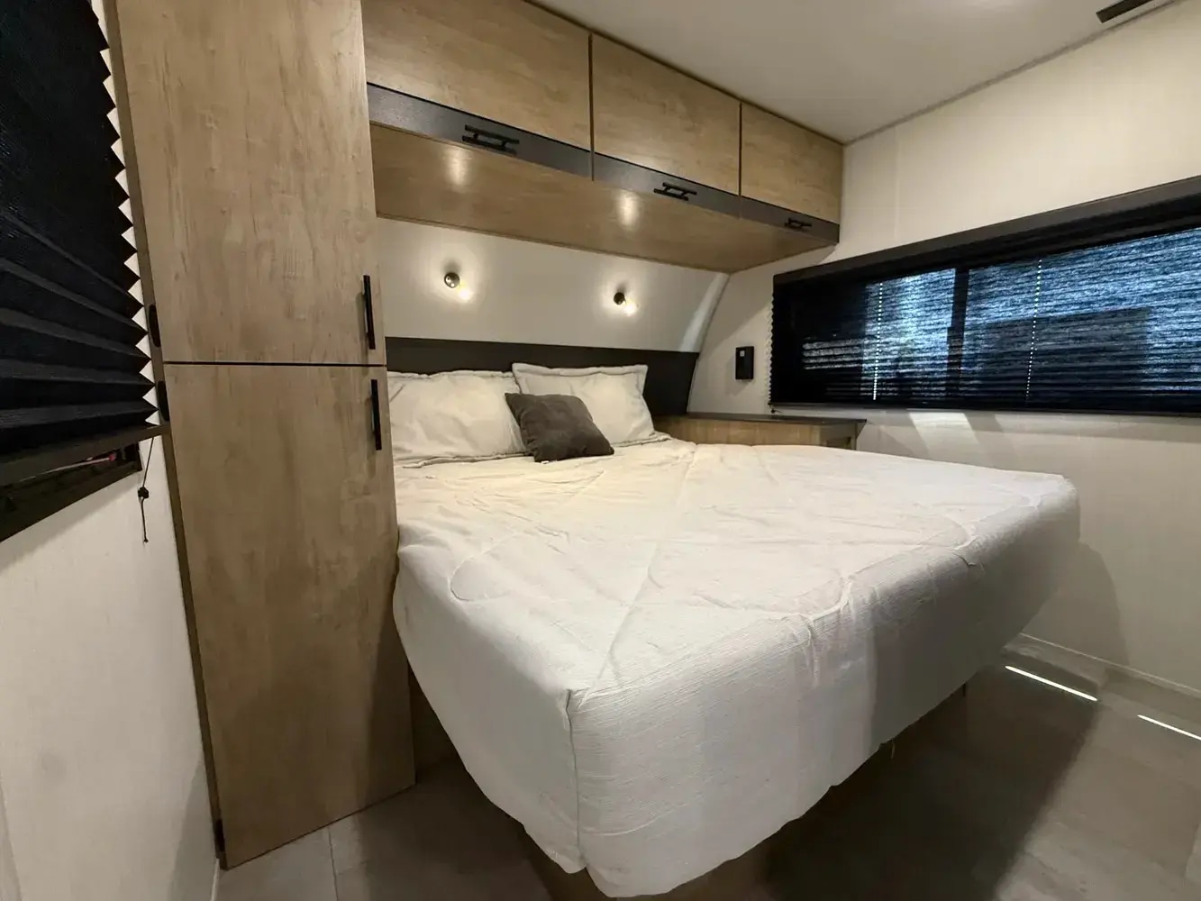 RV rental bedroom: a white-covered bed, wall-mounted lights, overhead storage, and a window with dark blinds.. Forest River Campsite Reserve 2026