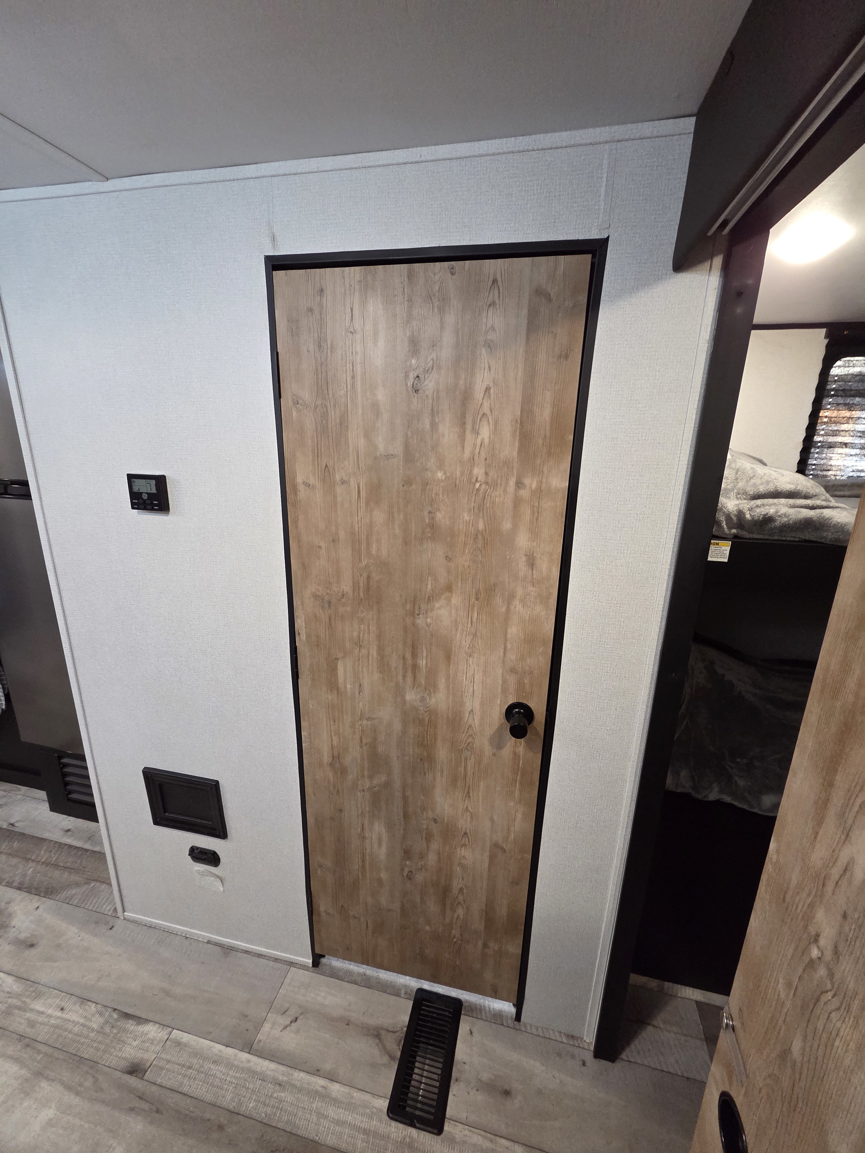 Modern RV rental: a wood-look door, textured grey walls with thermostat, floor vent, and a glimpse of a bunk.. Keystone RV Springdale 2022