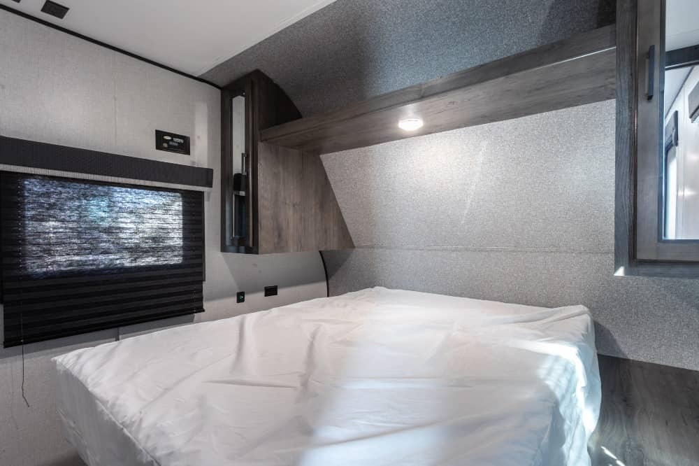 Cozy RV rental bedroom showcasing a white-covered bed, window with dark blinds, overhead shelf, and side cabinets.. Starcraft Autumn Ridge 2022