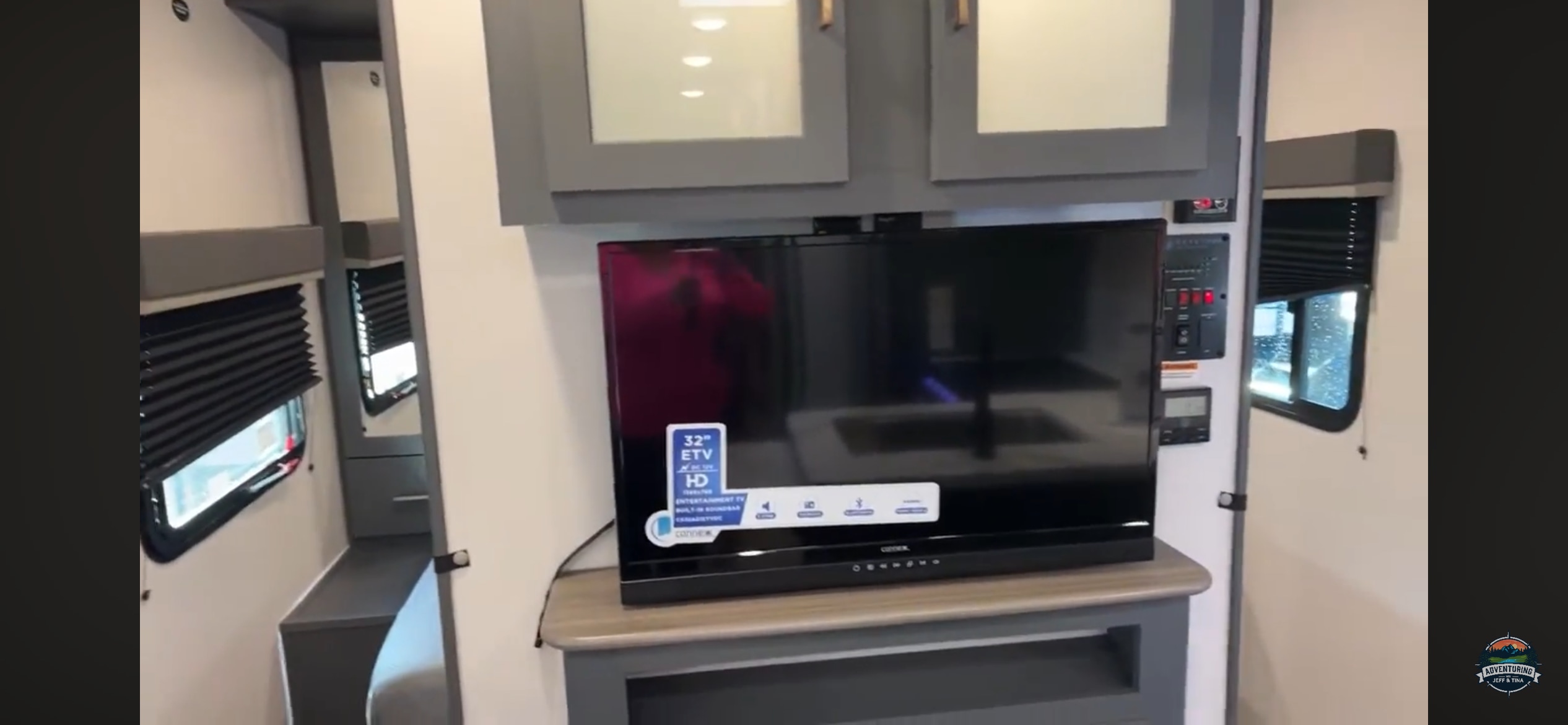 RV rental interior: A TV is centered under gray cabinets, next to control panels and windows with blinds, highlighting a compact living space.. Keystone RV Bullet Ultra Lite 2023