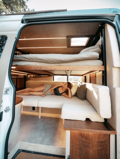 Cozy RV rental interior shows a woman relaxing on a sofa, with a comfy bunk bed above. Ideal for your next van adventure.. Other Sandy Vans 2025