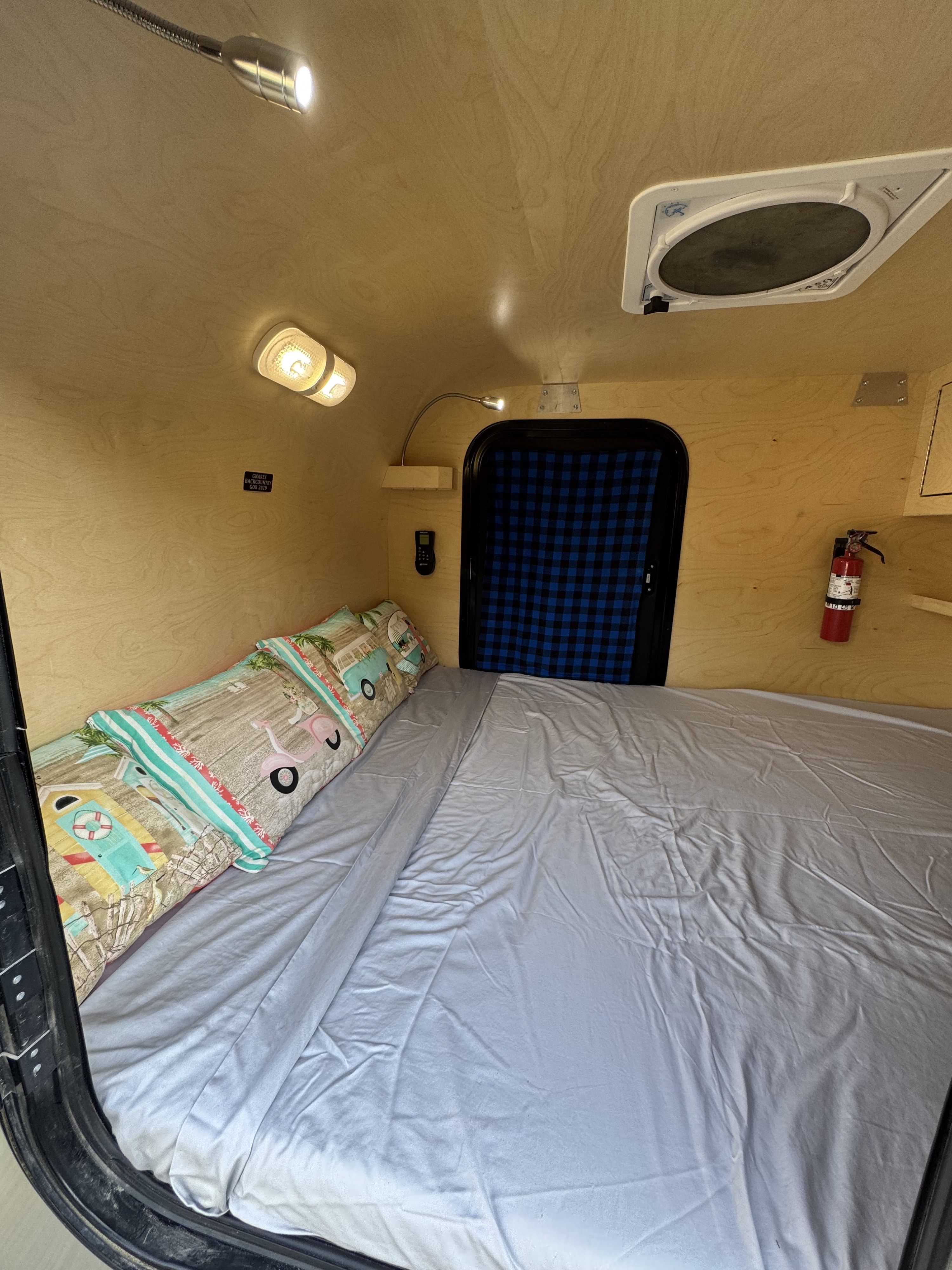 Inside a compact rv rental: bright wooden walls, a comfy bed with grey sheets & themed pillows, lights, ceiling vent, and safety gear.. Gnome Homes Inc. Backcountry 2020