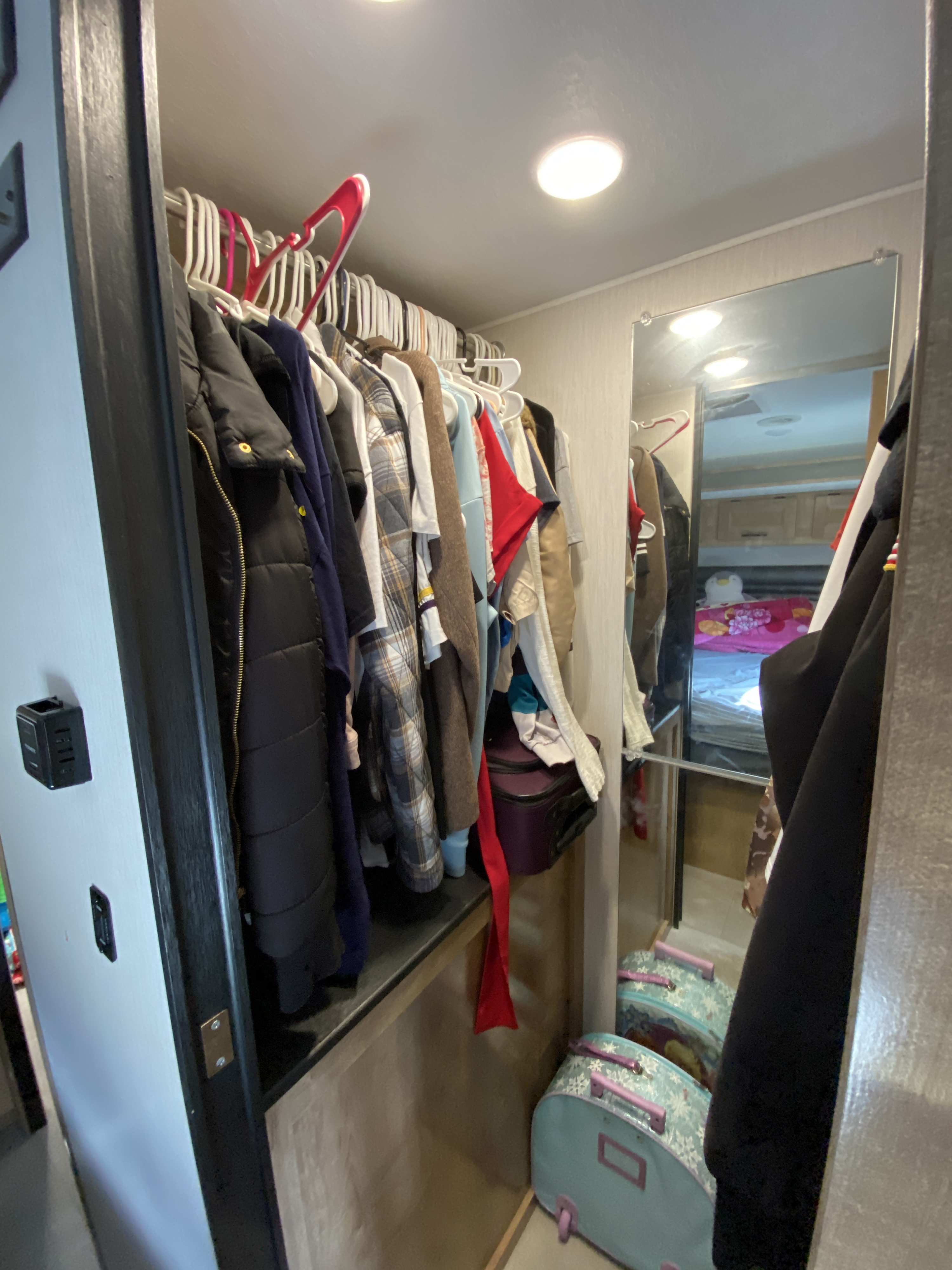 Inside an RV rental, a compact closet filled with hanging clothes, small suitcases, and a mirror reflecting the bed.. Coachmen Leprechaun 311fs 2020
