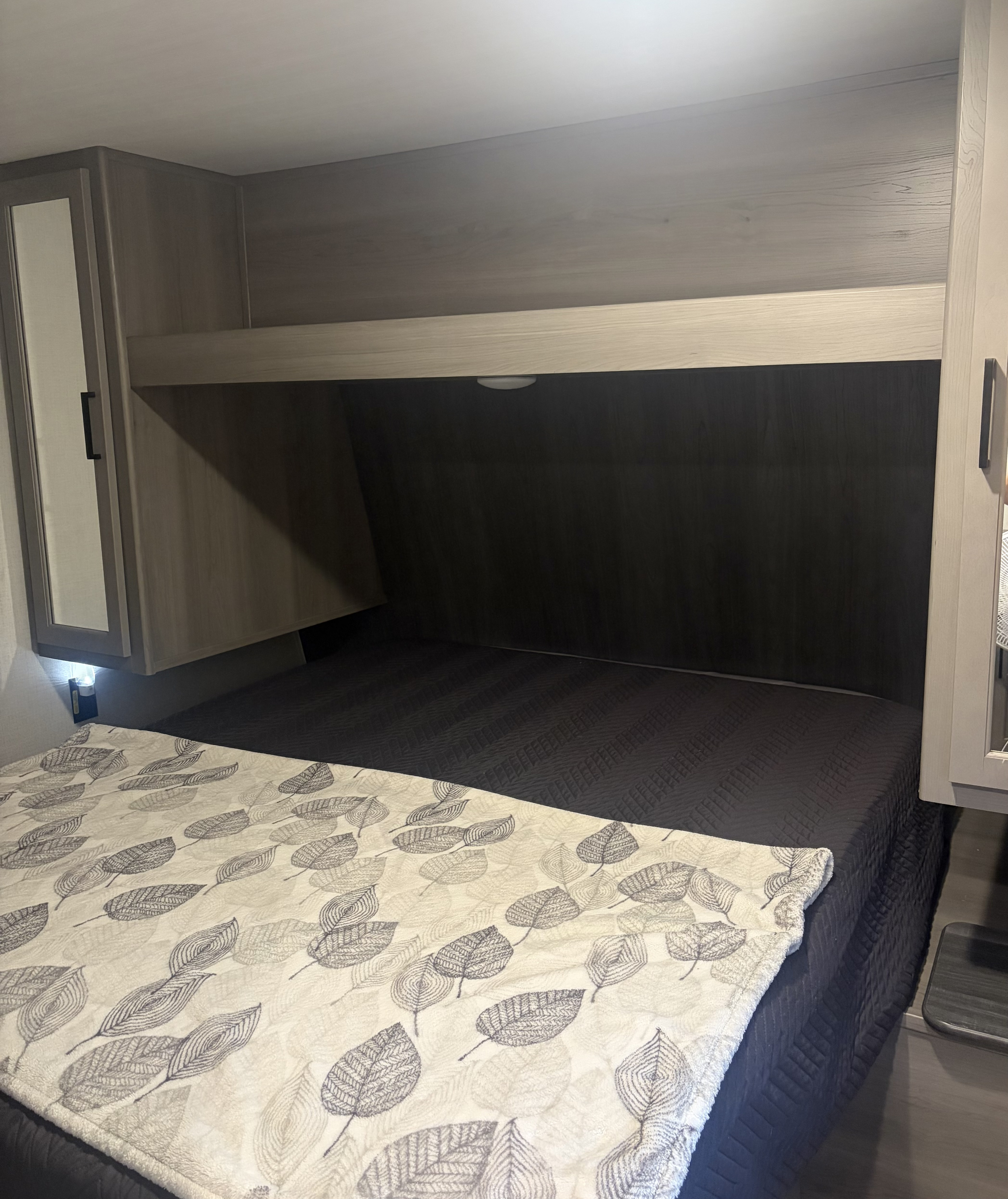 Cozy RV rental bedroom featuring a dark quilted bed, leaf-patterned blanket, and integrated overhead storage with lighting.. Keystone RV Hideout 2024