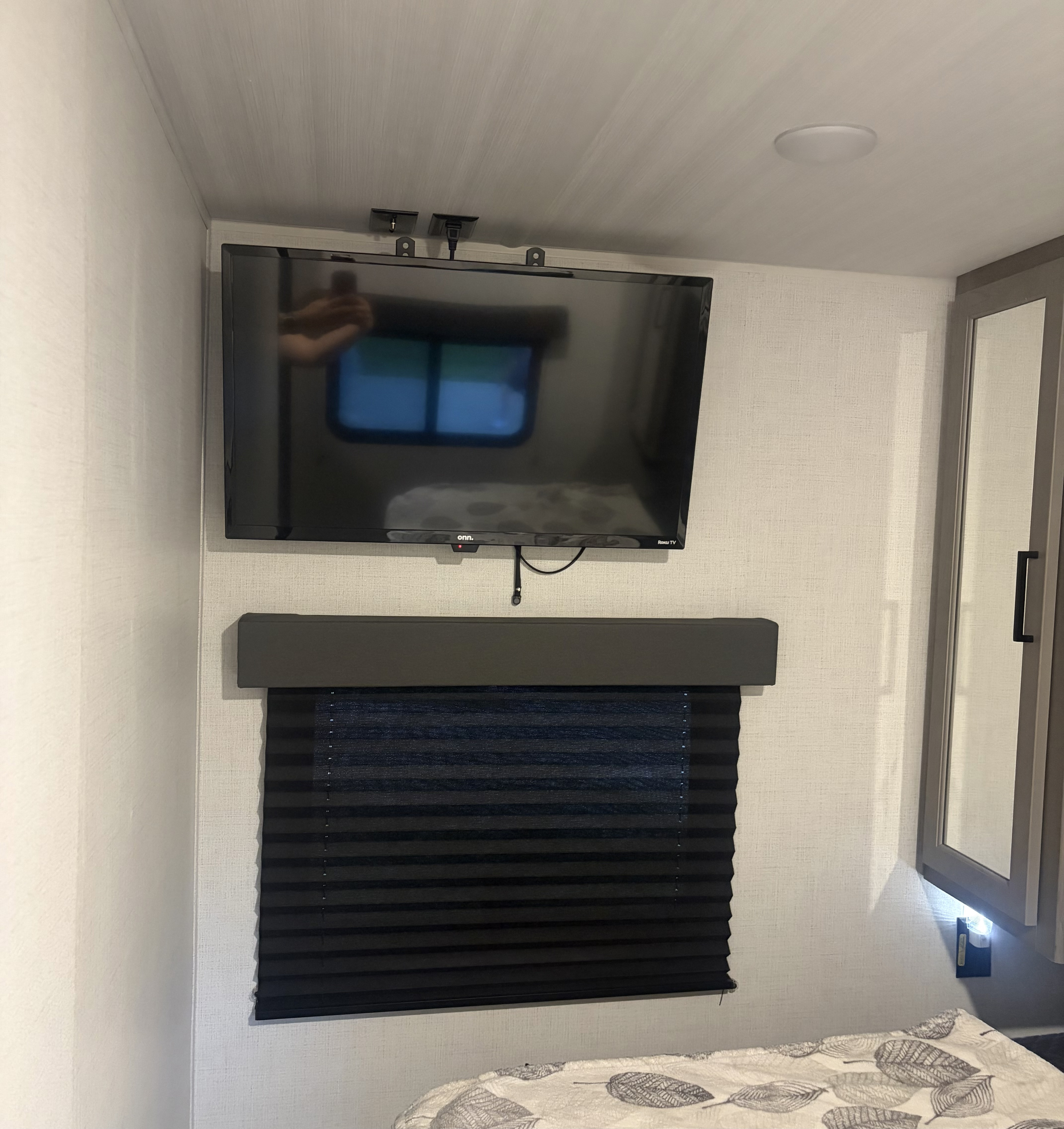 Inside an RV rental, a compact bedroom shows a wall-mounted TV above a window with a dark pleated blind and soft bedding.. Keystone RV Hideout 2024