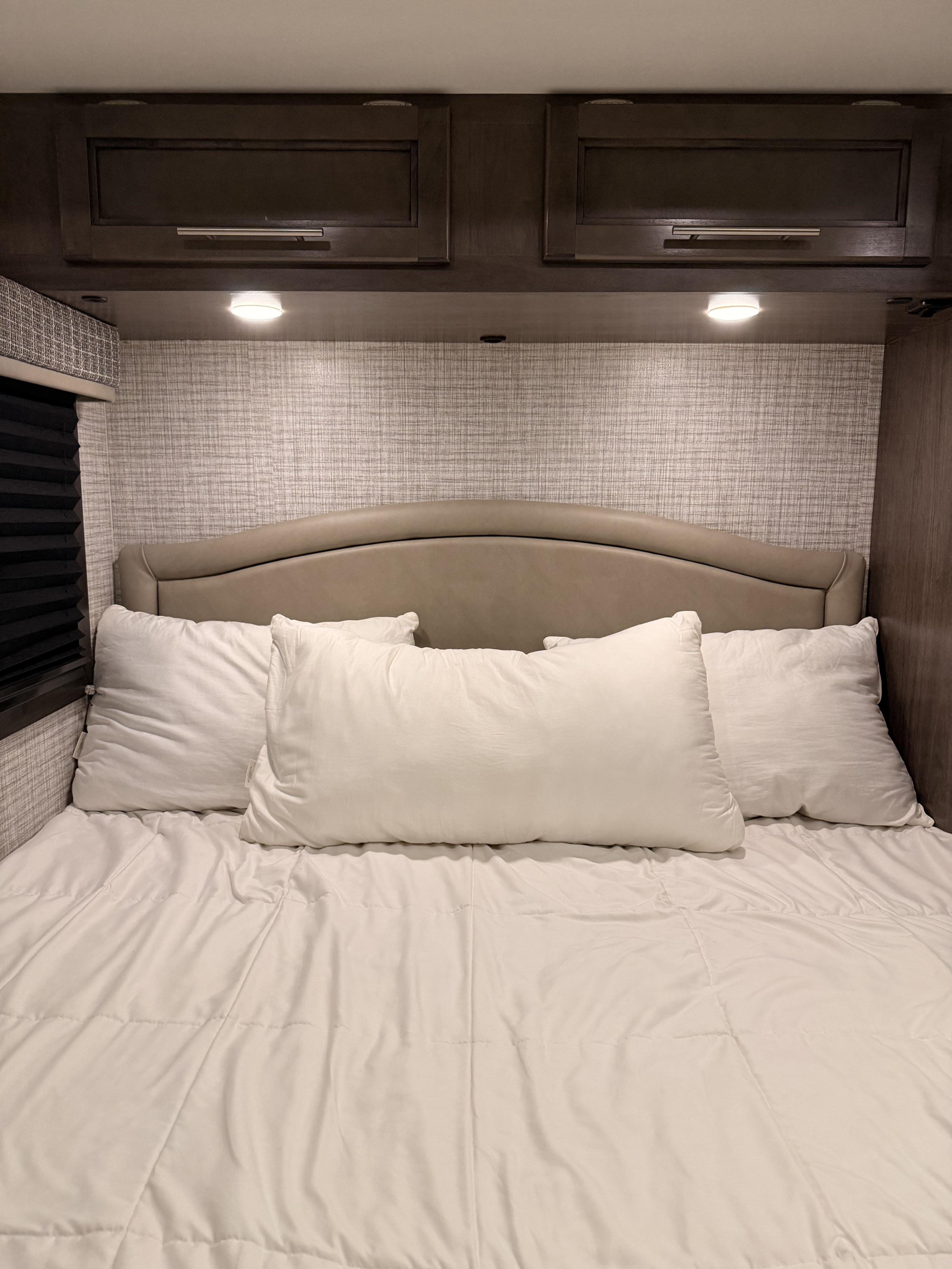 Cozy RV rental bedroom featuring a bed with white pillows and comforter, dark wood cabinets, and task lighting.. Jayco Melbourne 2024