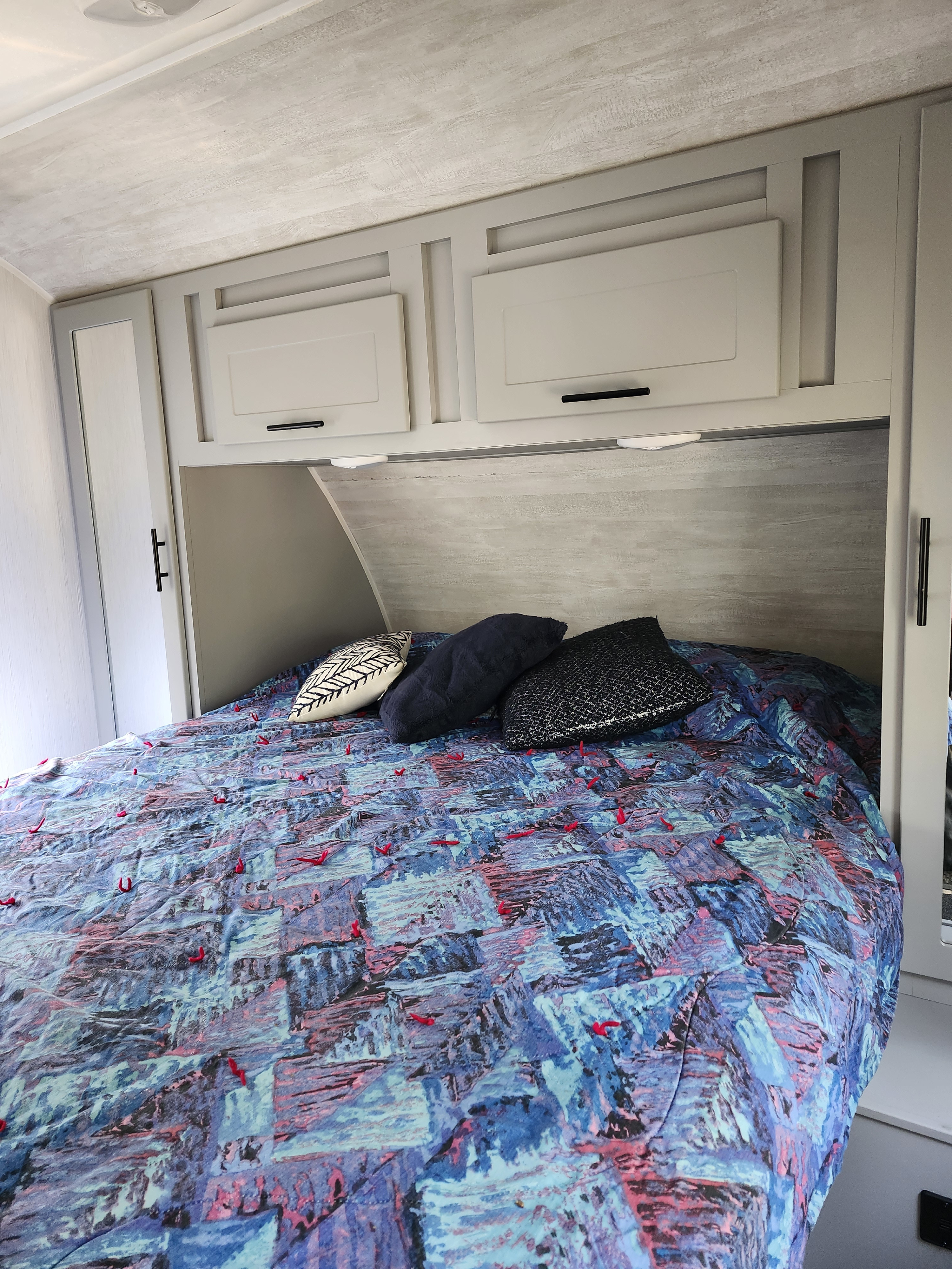 This RV rental bedroom features a bed with a vibrant blue and red patterned comforter, pillows, and light overhead storage cabinets.. Forest River Vibe 2023