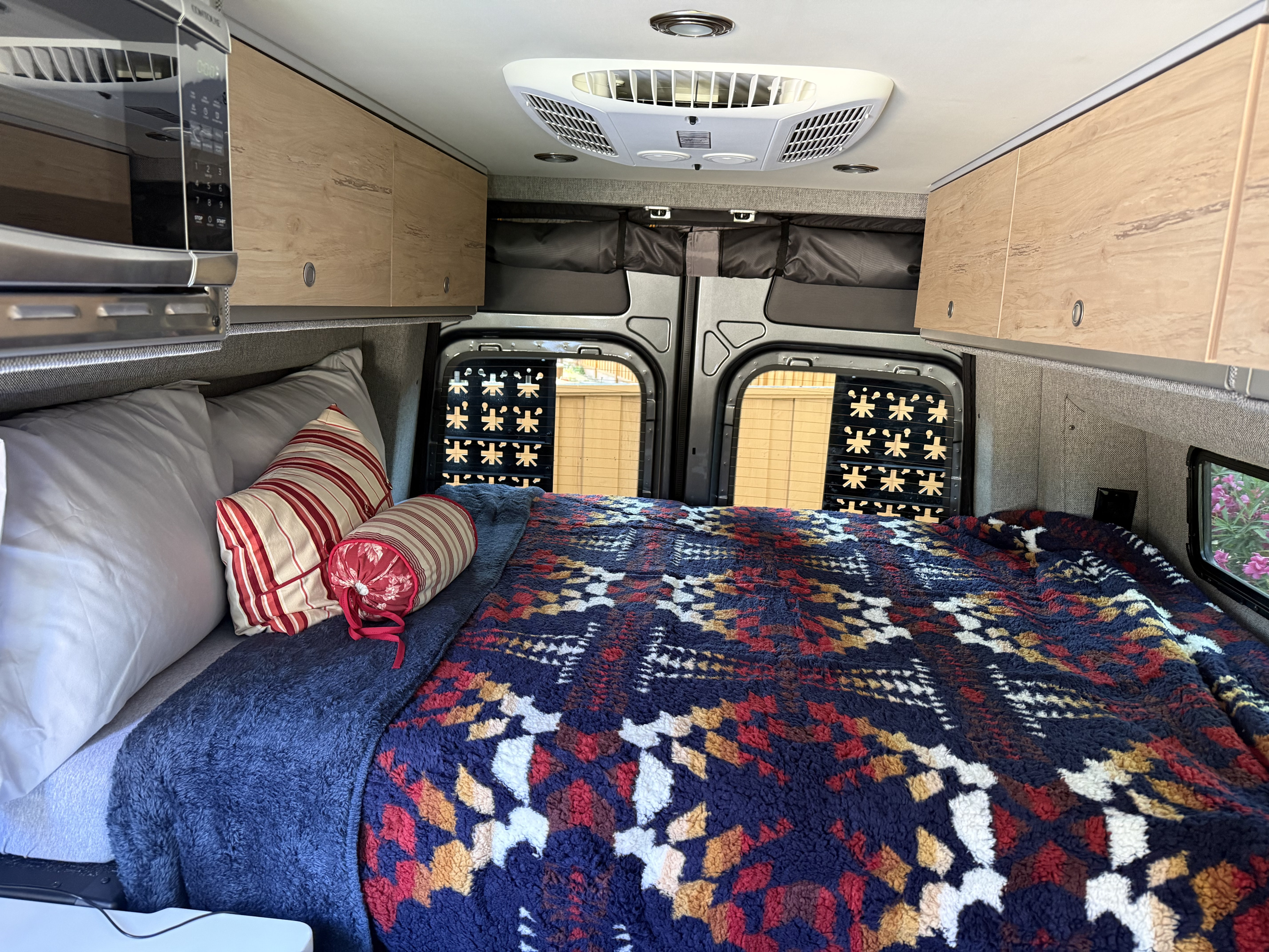 Cozy RV rental bedroom with microwave, overhead storage, AC unit, and patterned bedding. Perfect for comfortable van life.. Mercedes Sprinter Motorhome Campervan RV 2025