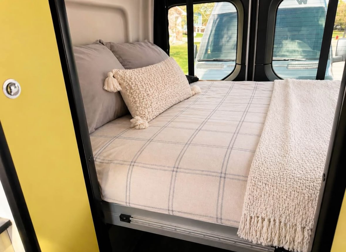 Inside an RV rental: a neatly made bed with plaid sheets, grey pillows, and a textured throw, viewed from the foot.. Winnebago Revel 2020