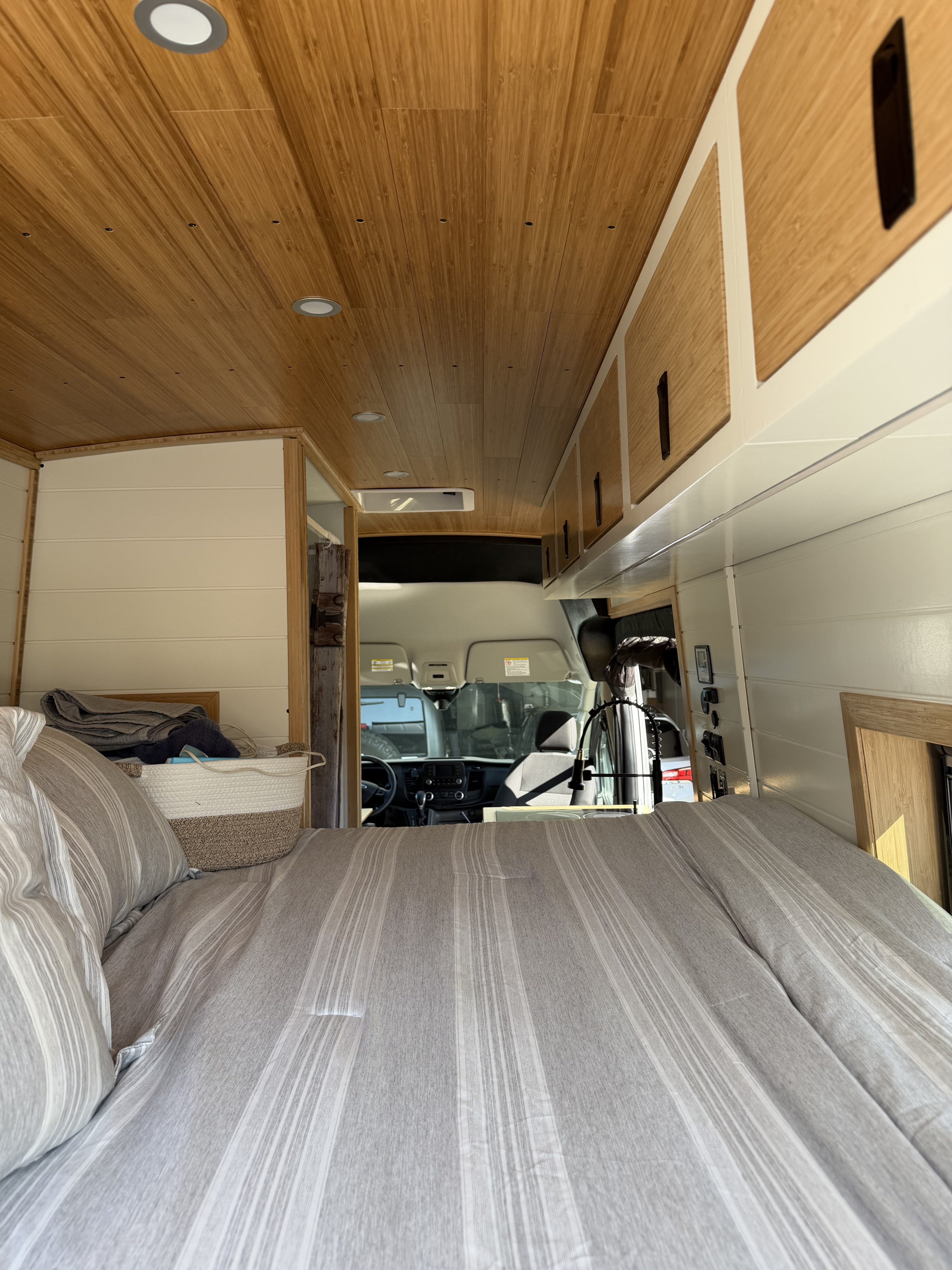 Cozy RV rental interior with a striped bed, wood ceiling, and a clear view to the driver's cabin in a camper van.. Ford Custom Transit 2025