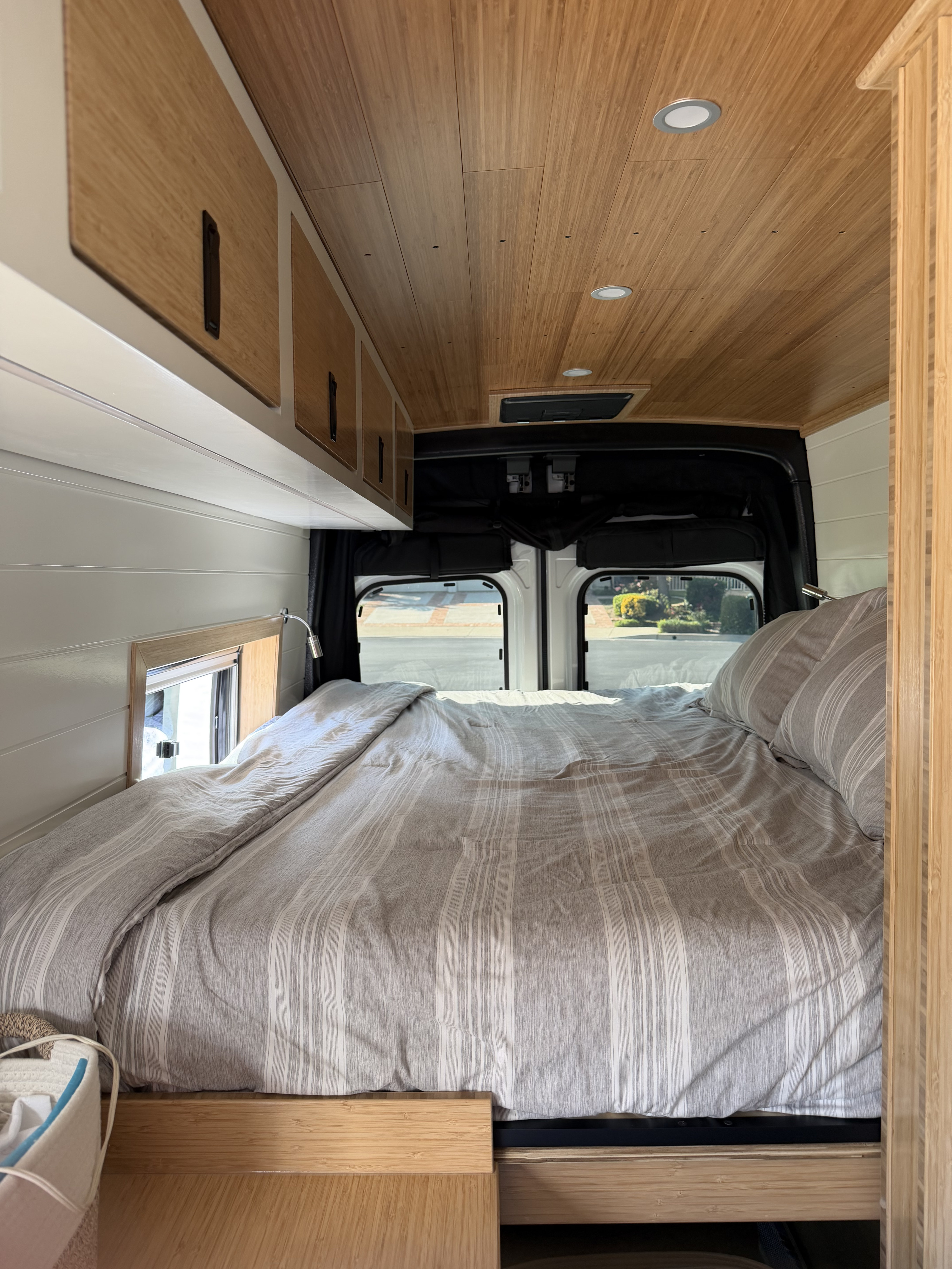 RV rental bedroom interior: cozy bed with striped linens, bamboo ceiling & cabinets, white walls, and rear windows.. Ford Custom Transit 2025