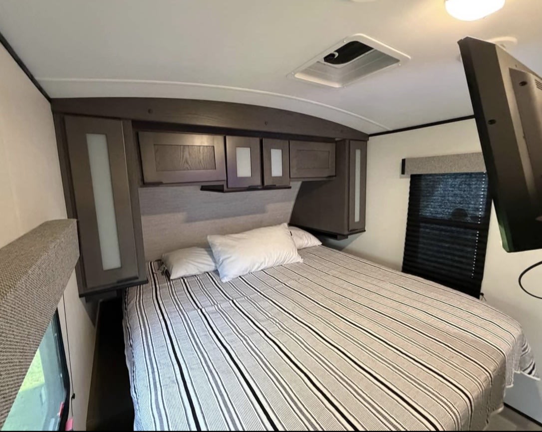 Cozy rv rental bedroom with a striped bed, overhead cabinets, and a wall-mounted TV.. Cruiser RV MPG Ultra Lite 2021