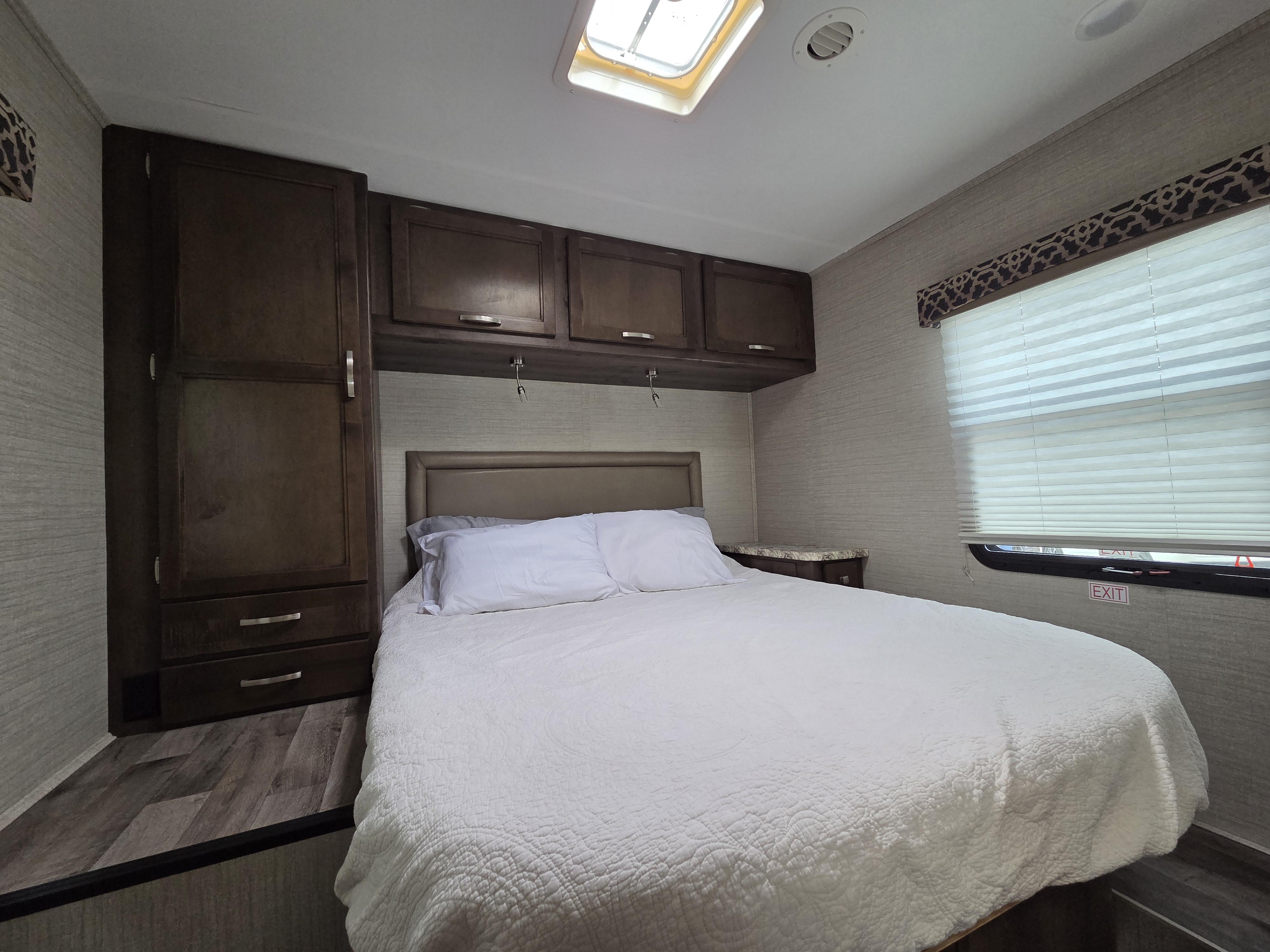 This RV rental features a cozy bedroom with white bedding, dark wood cabinets, and a window with blinds & an exit sign.. Jayco Redhawk 2021