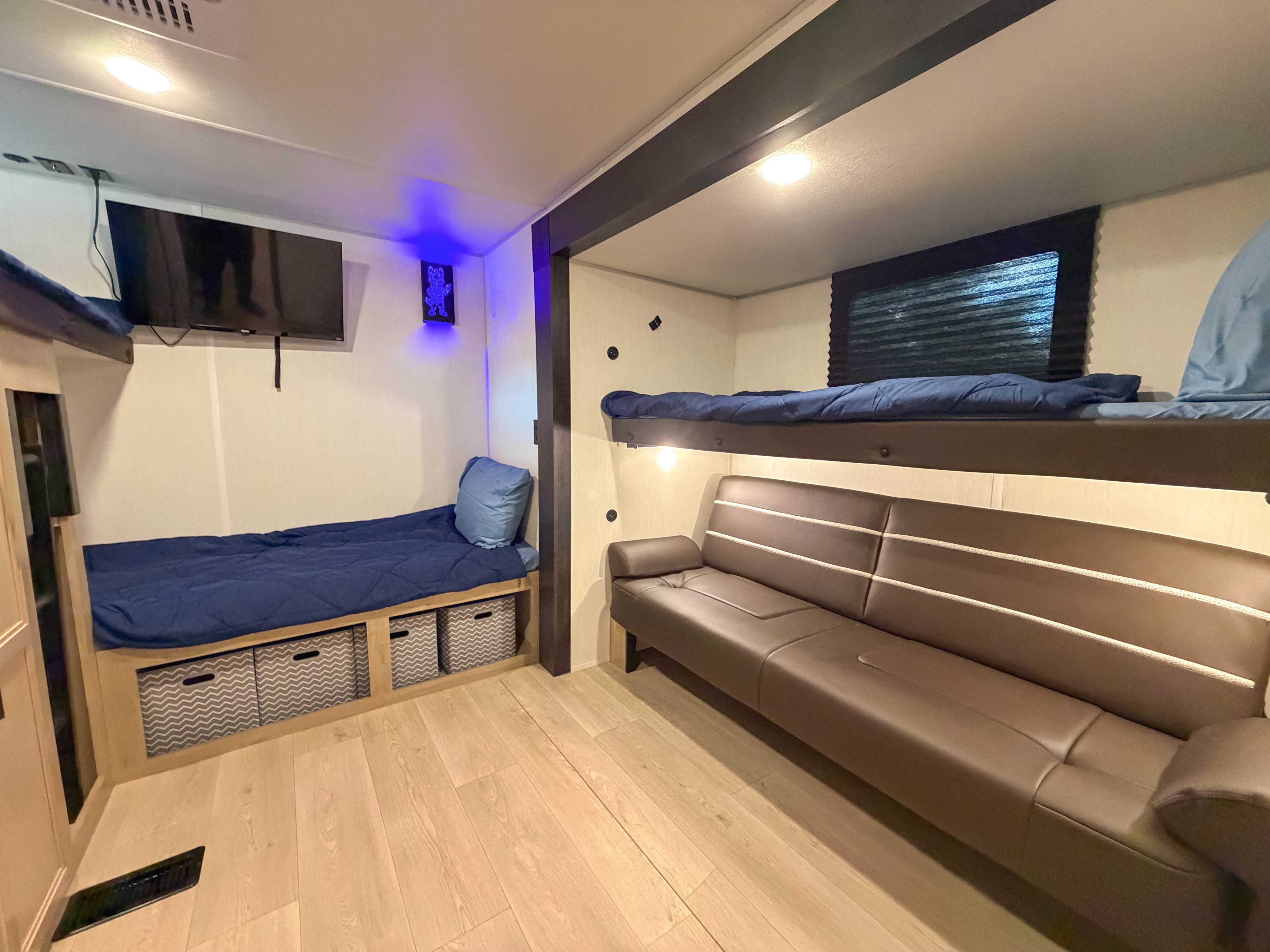 Cozy RV rental bunk room with light wood floors, blue bunks, brown sofa, wall-mounted TV, and convenient storage.. Forest River CHEROKEE WOLF DEN 301BH 2026