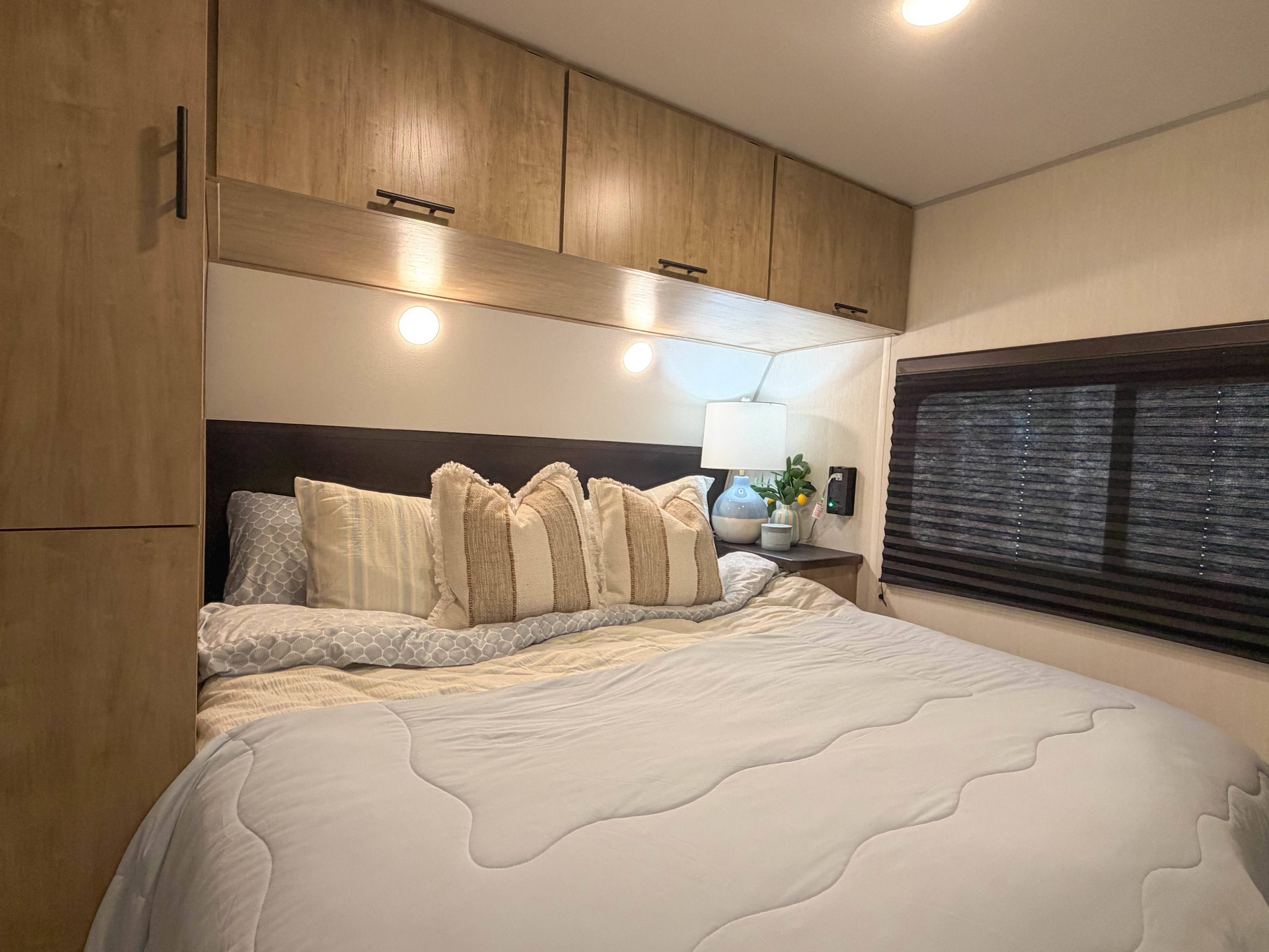 Relax in this cozy RV rental bedroom featuring a comfortable bed with patterned pillows, overhead storage, and a nightstand with a lamp.. Forest River CHEROKEE WOLF DEN 301BH 2026
