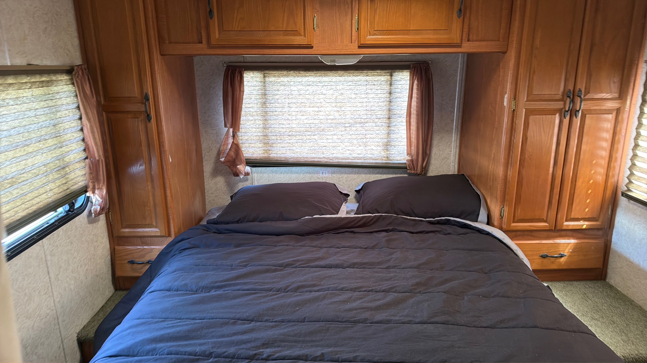 An RV rental bedroom with a neatly made bed, dark bedding, a window with closed blinds, and built-in wooden cabinets.. Coachmen Leprechaun 2003