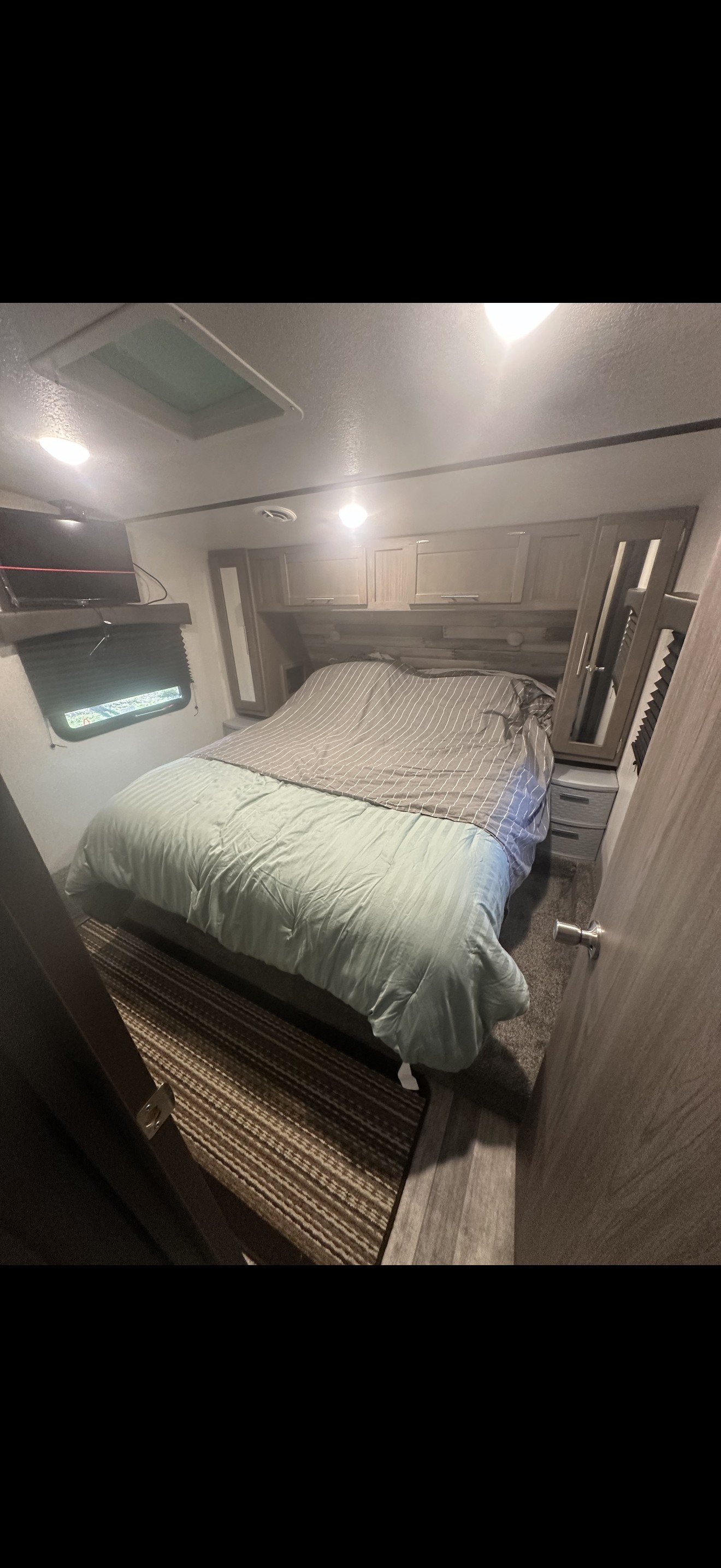 RV rental bedroom: a cozy bed with striped comforter, overhead cabinets, a small TV, window, and ceiling skylight.. Cross Roads RV 4Ydfvlr25n6360061 2022