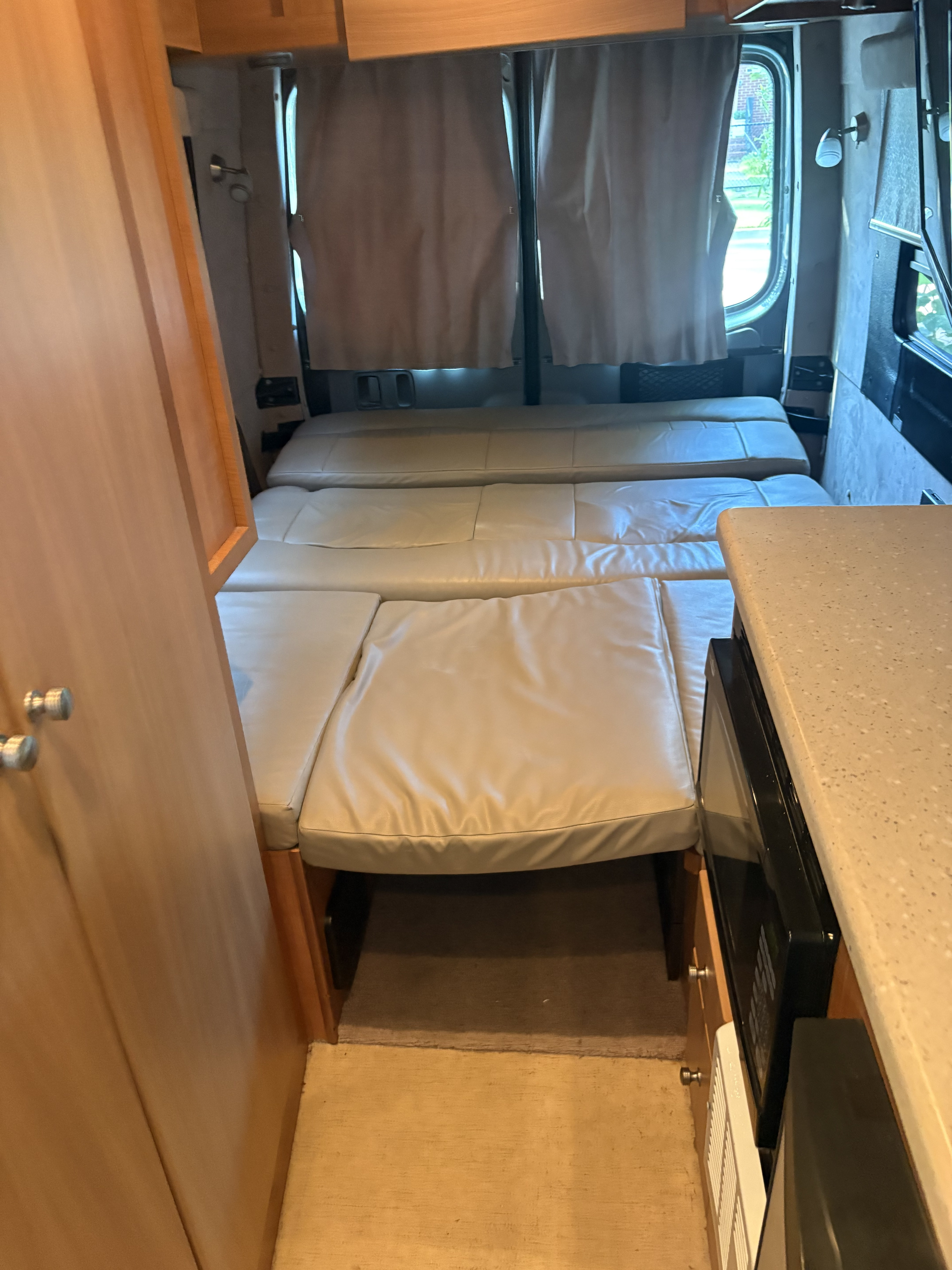 Compact RV rental interior with a light-colored, convertible bed/seating area, wooden cabinetry, and a kitchenette with a microwave.. Winnebago Eurovan Camper 2010 era limited 2010