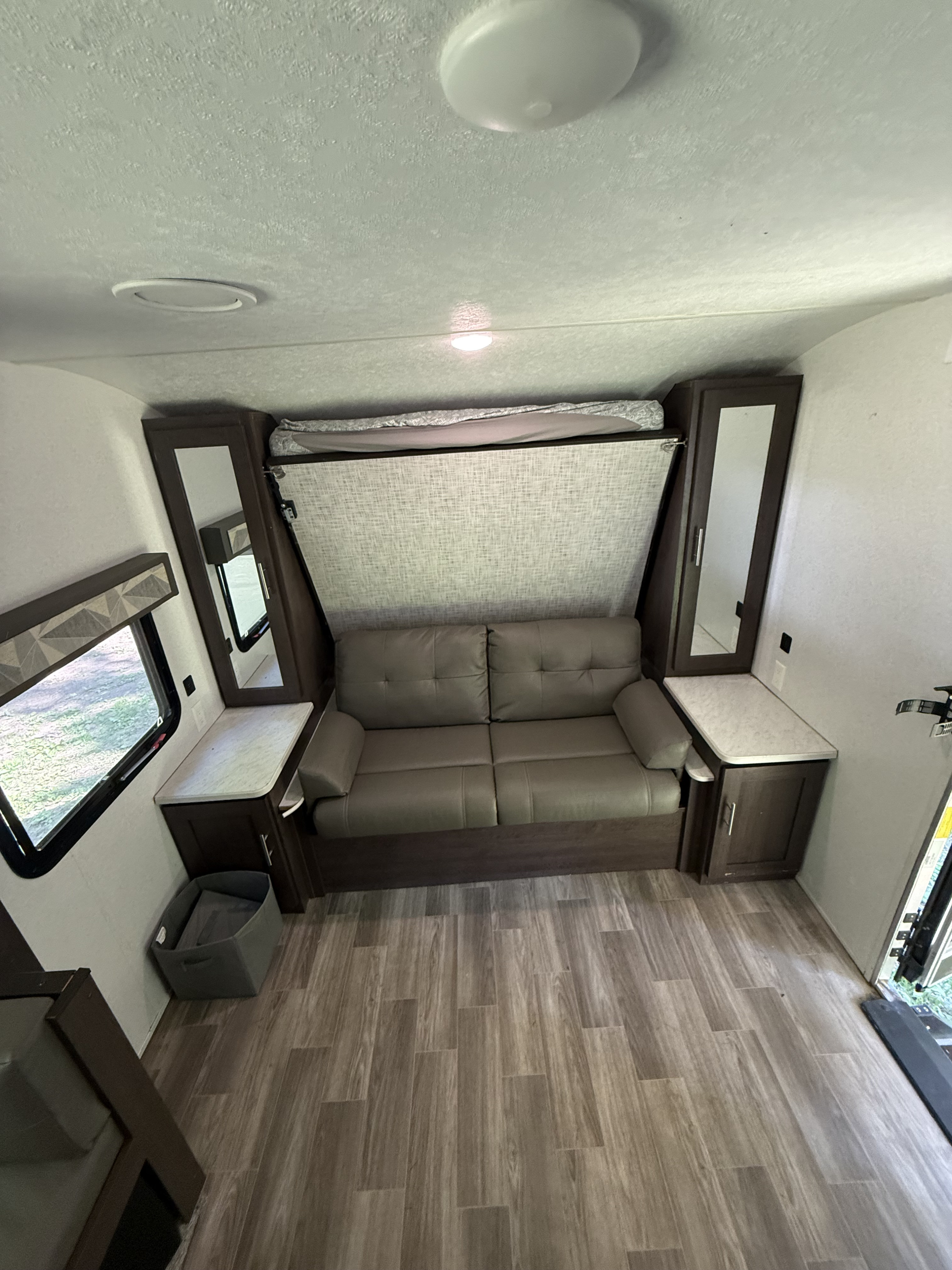 RV rental living area with a gray sofa converting to a wall bed. Features wood-look floor, mirrored cabinets, and windows.. Forest River Wildwood X-Lite 2019