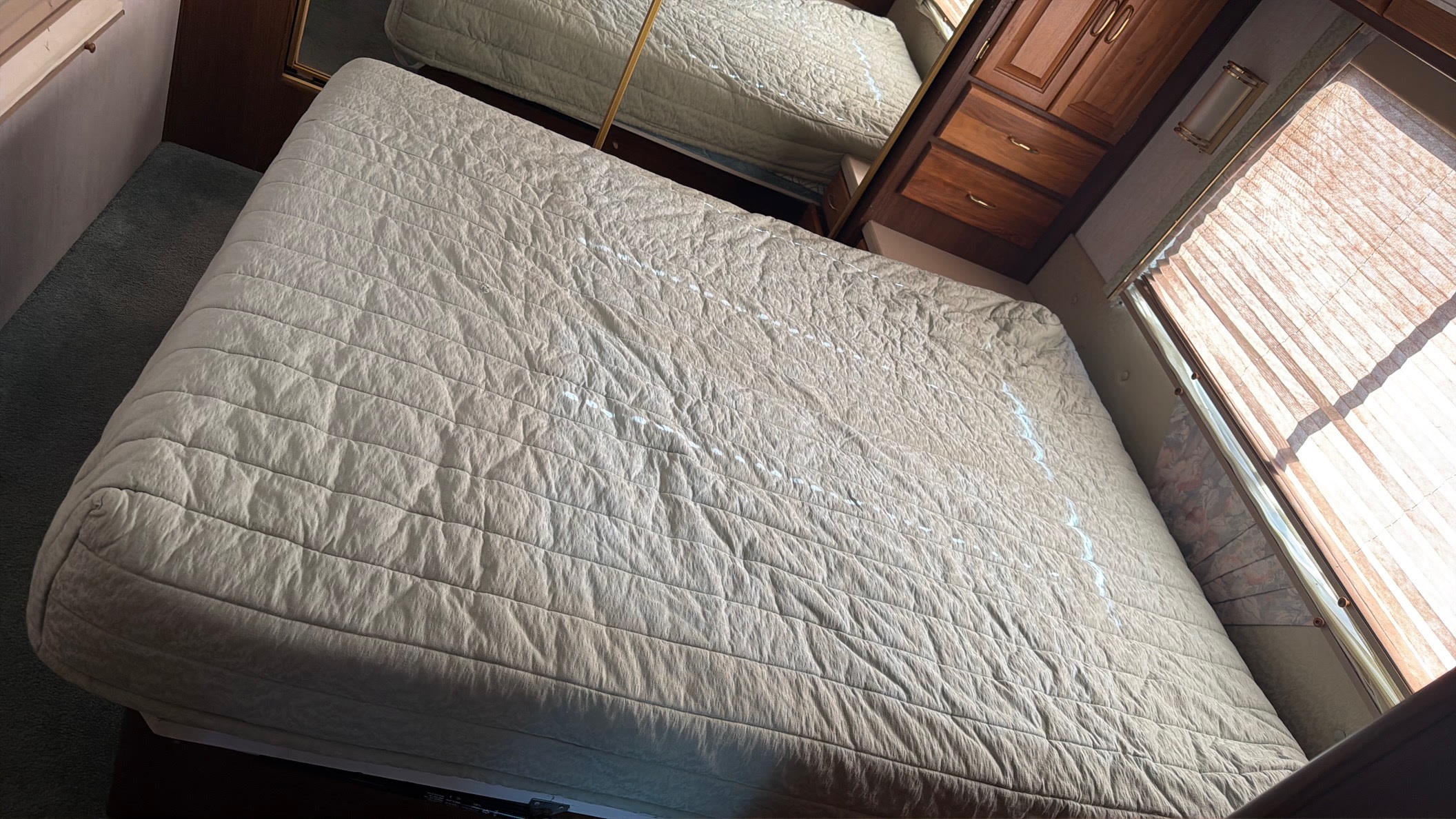 Inside an RV rental, showing a bed with quilted bedding, wood cabinets, and a sunlit window with blinds.. Intermountain Rv TROPICAL 1997