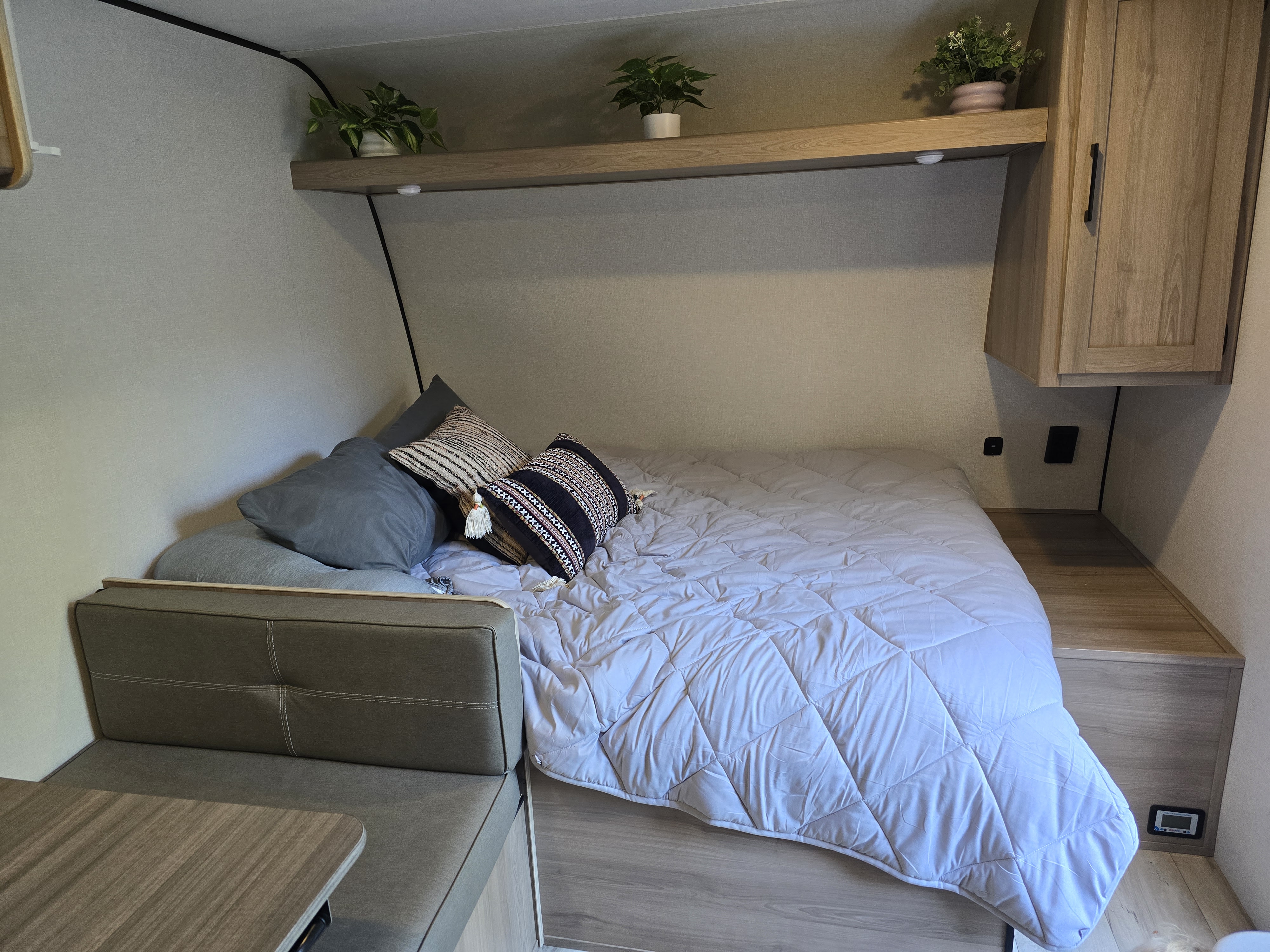 This RV rental features a cozy bed with pillows, a dinette, and overhead shelf with plants in a compact camper interior.. Keystone RV Coleman Light 2025