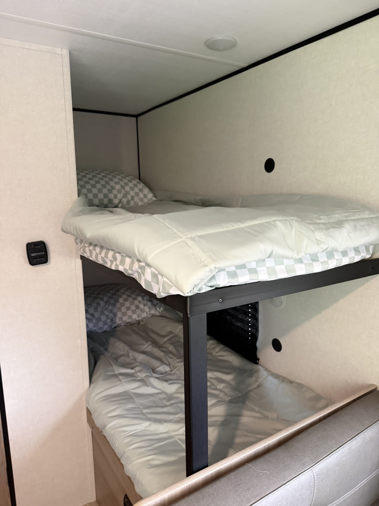 Inside an RV rental, two neatly made bunk beds with checkered bedding and light green comforters. A control panel is on the left.. Keystone RV Coleman 2025