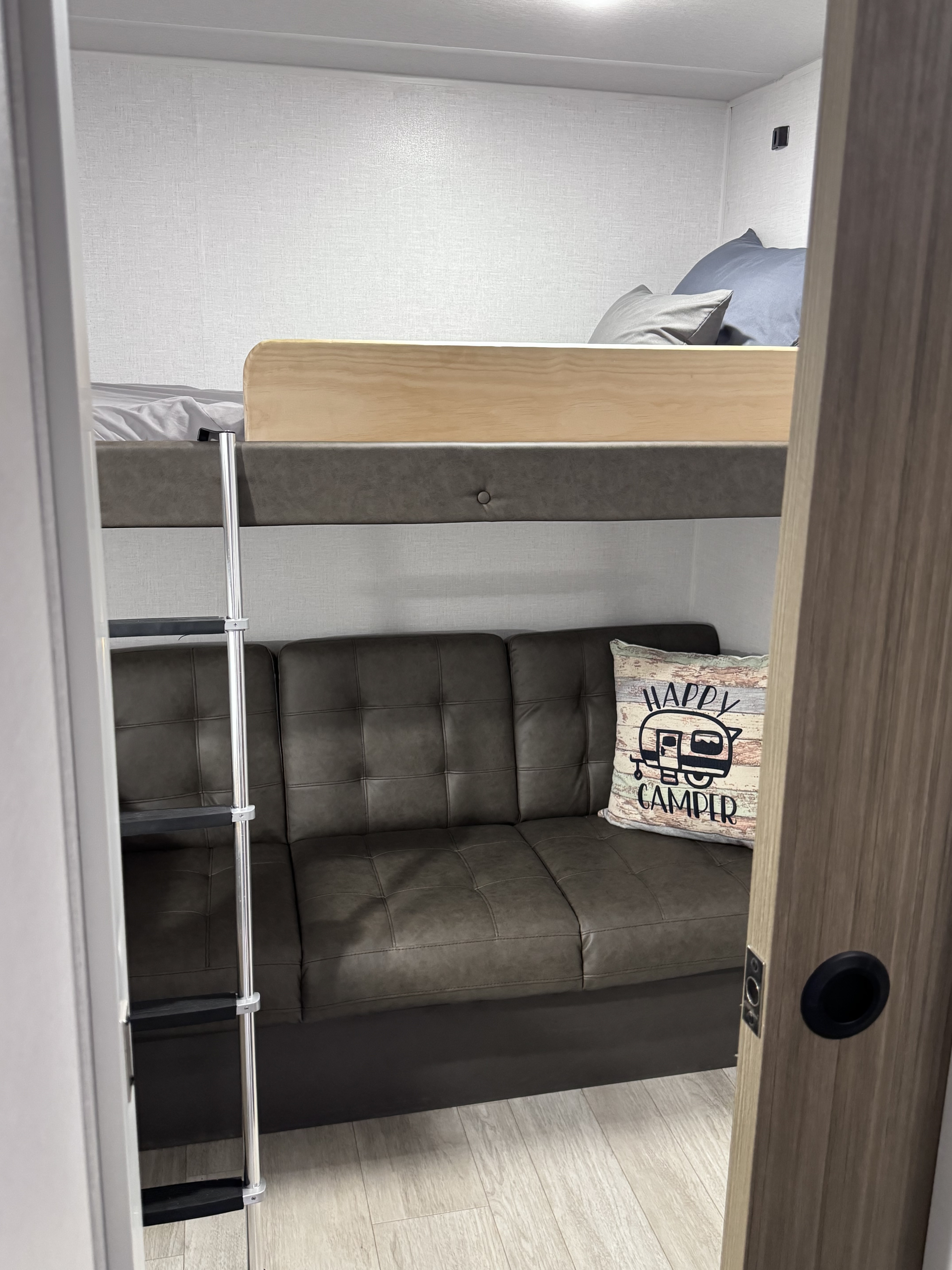 Cozy RV rental bunk area: top bed, ladder, and a dark gray sofa below with a 'Happy Camper' pillow.. Forest River X-Lite 2026