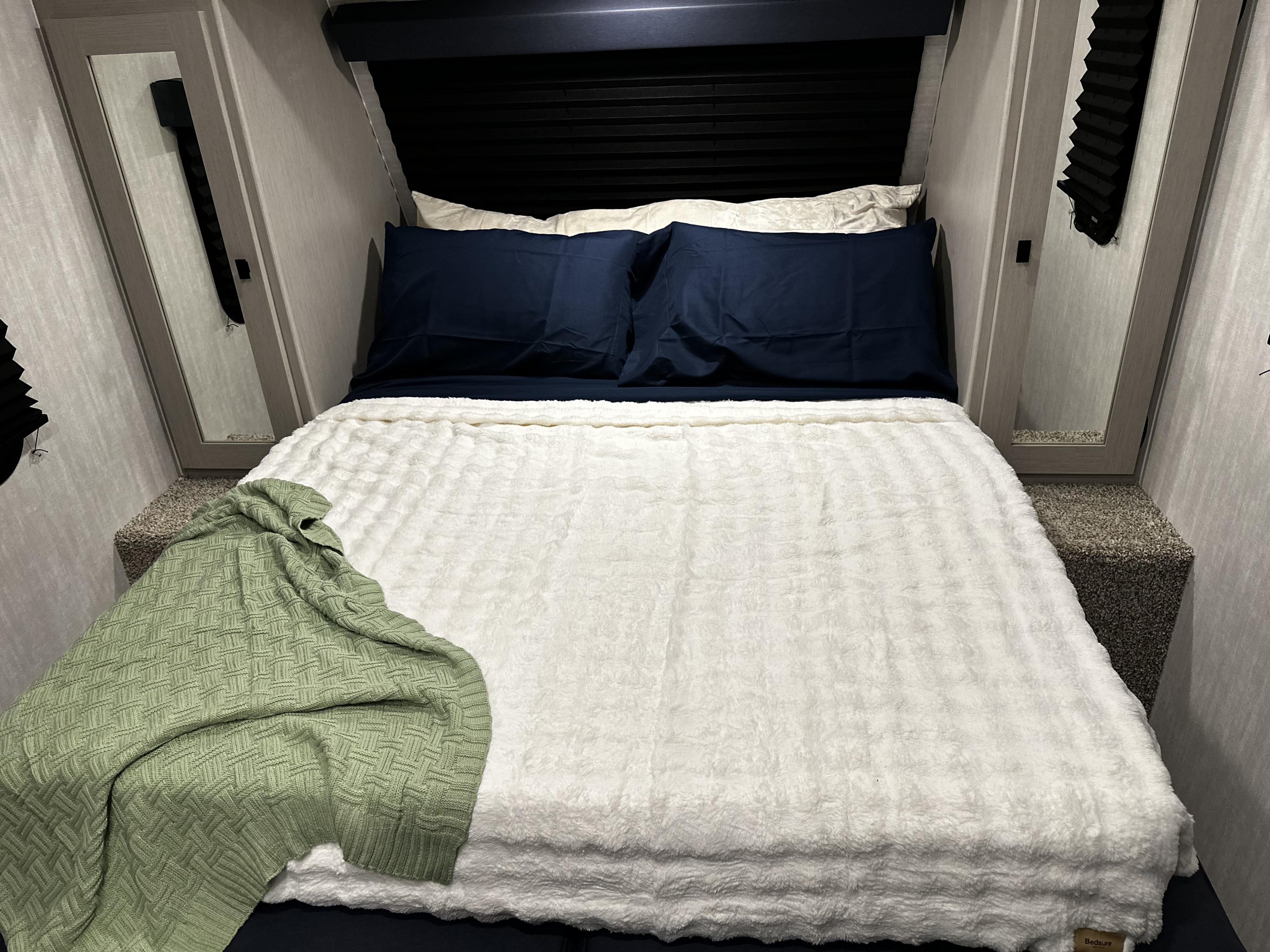Cozy RV rental bedroom with a neatly made bed, featuring navy sheets, a white fluffy blanket, and a green throw.. Coachmen RMT18R 2024