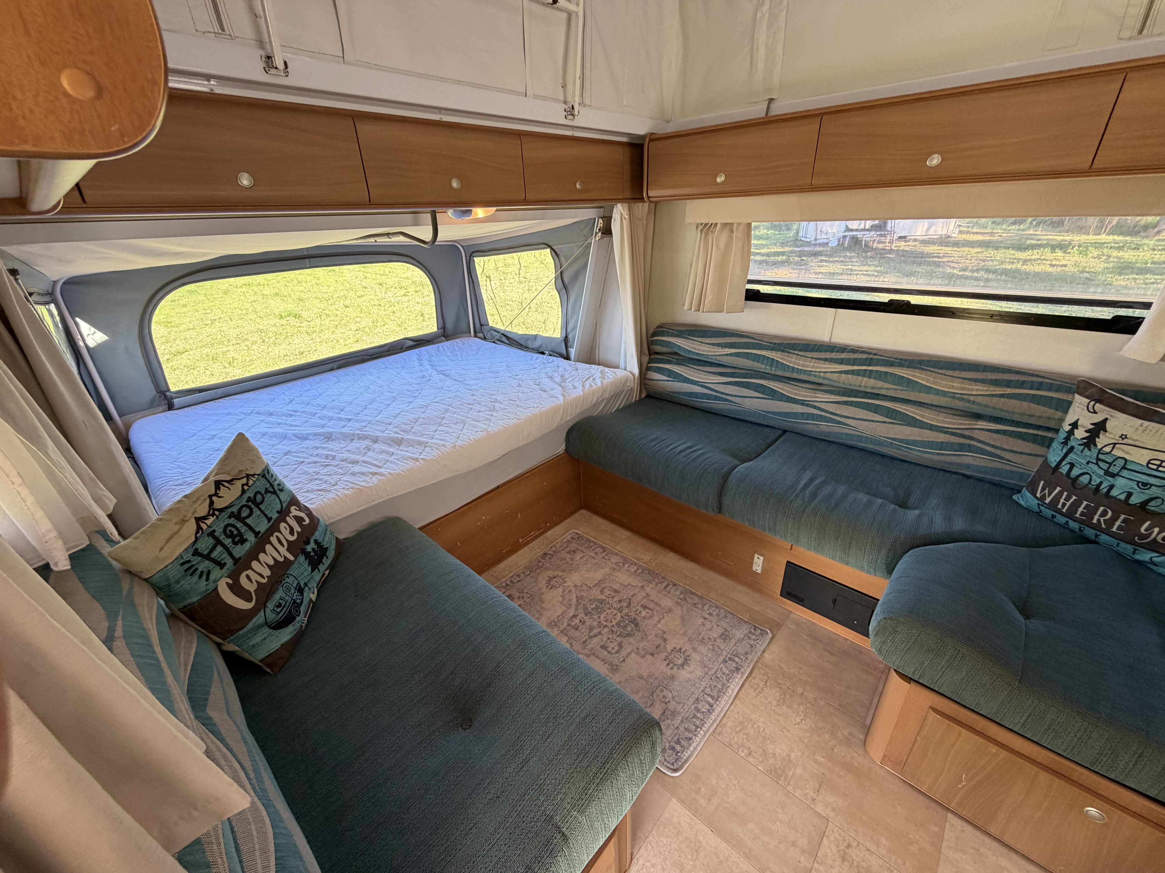 Inside a cozy RV rental: bed, teal seating with fun pillows, and wood overhead cabinets for storage.. jayco Expanda 2010