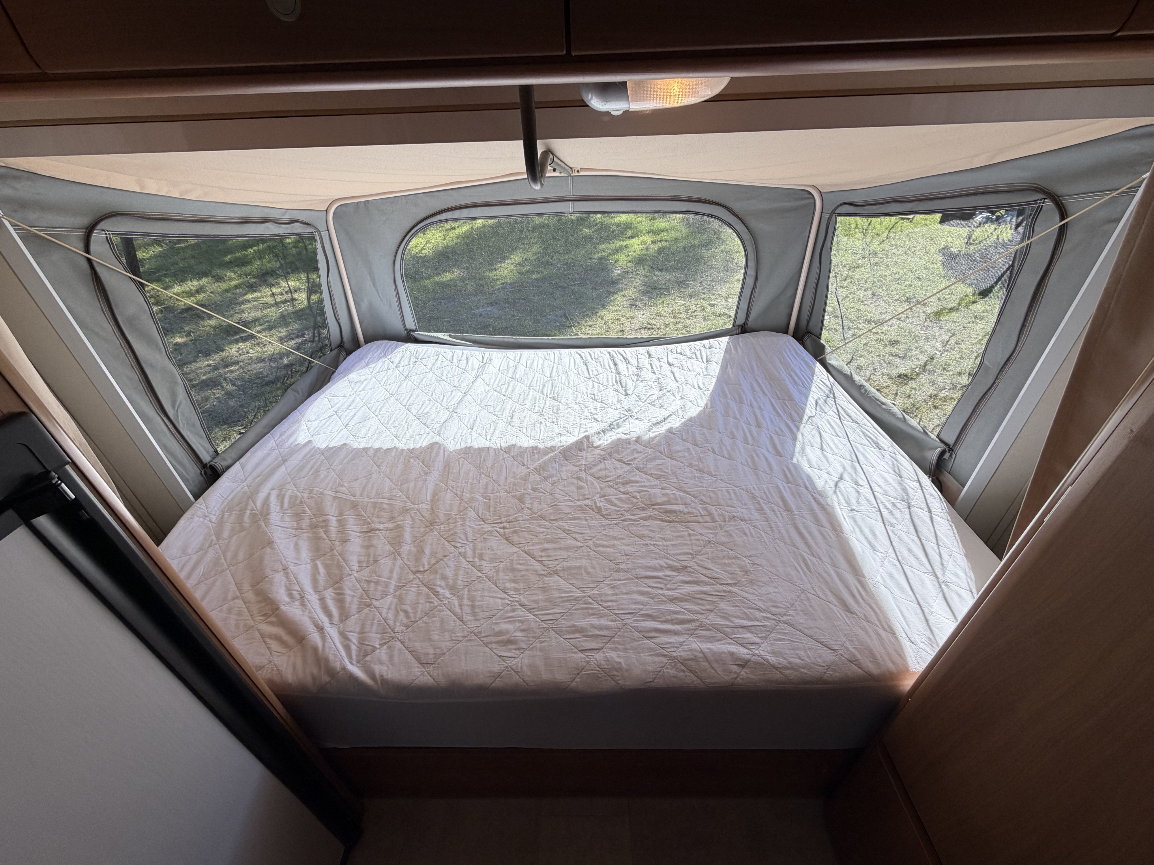 Cozy RV rental pop-up camper bed area with bright light and green outdoor views through surrounding windows.. jayco Expanda 2010