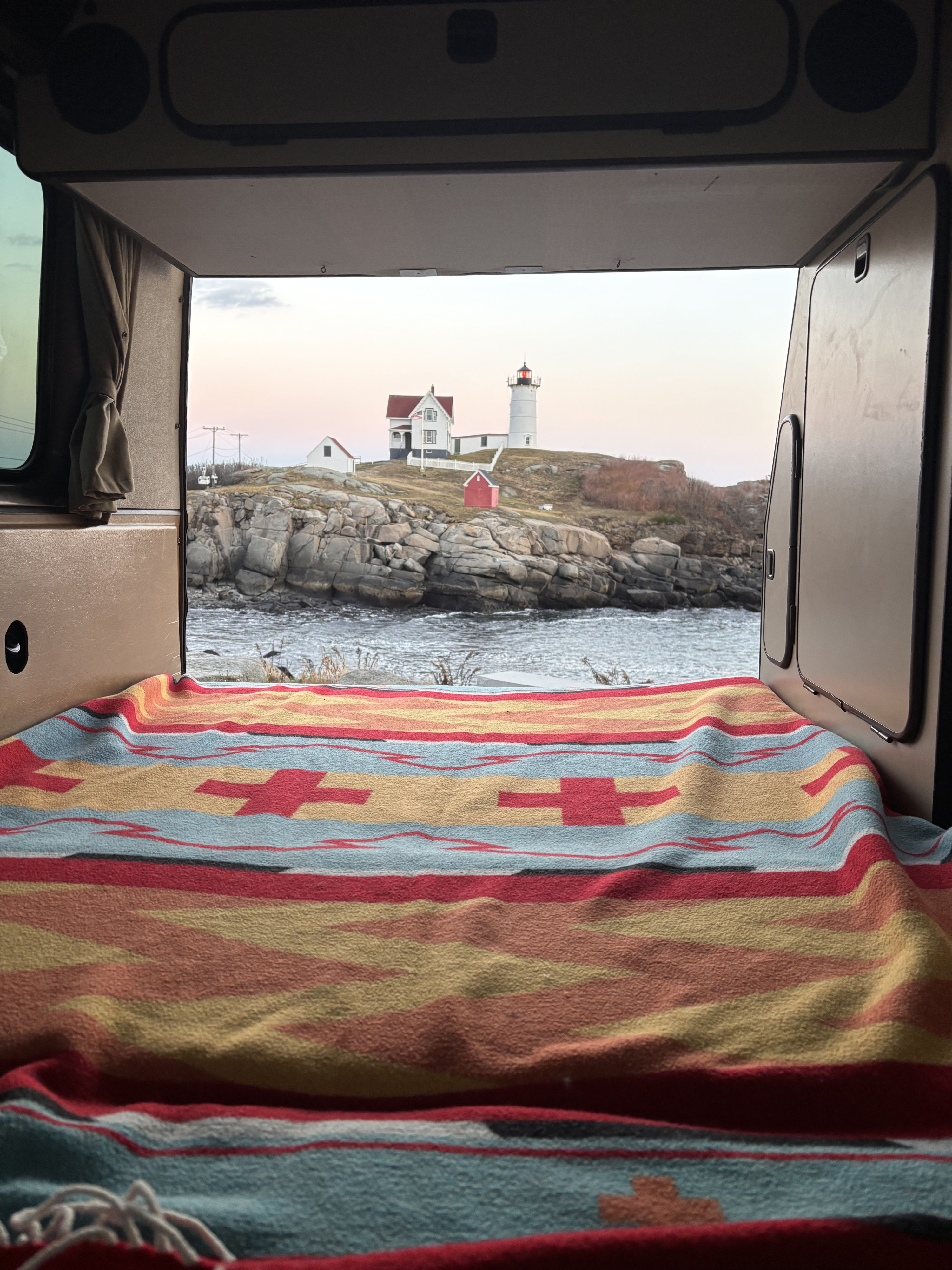 Cozy rv rental campervan bed with a vibrant blanket, offering a stunning ocean view of Nubble Lighthouse. Ideal for scenic travel.. Volkswagen Westfalia 1985