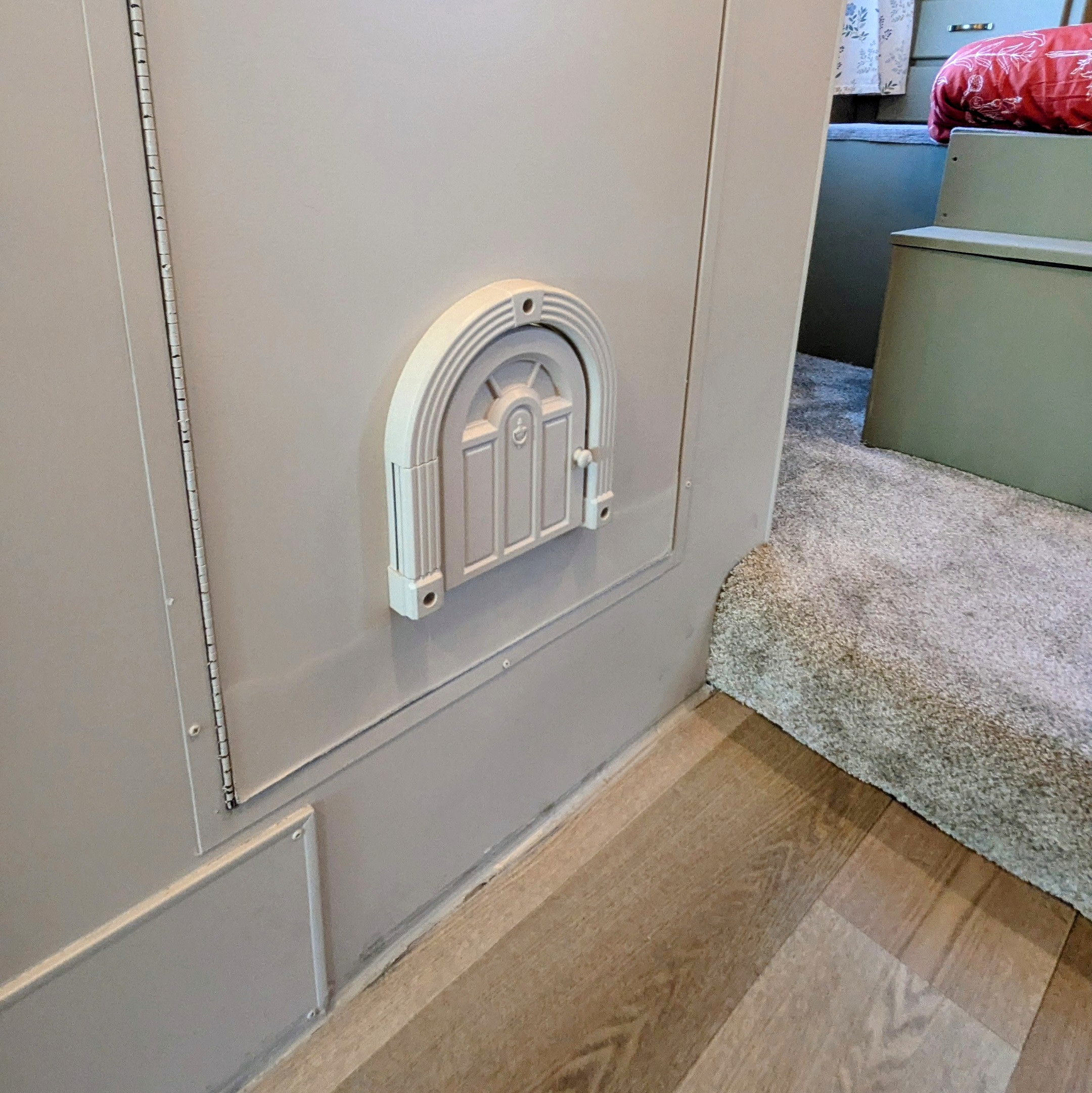 Inside an RV rental, a small, decorative arched access panel is on a light gray wall above wood flooring, transitioning to carpet.. Thor Majestic 2019