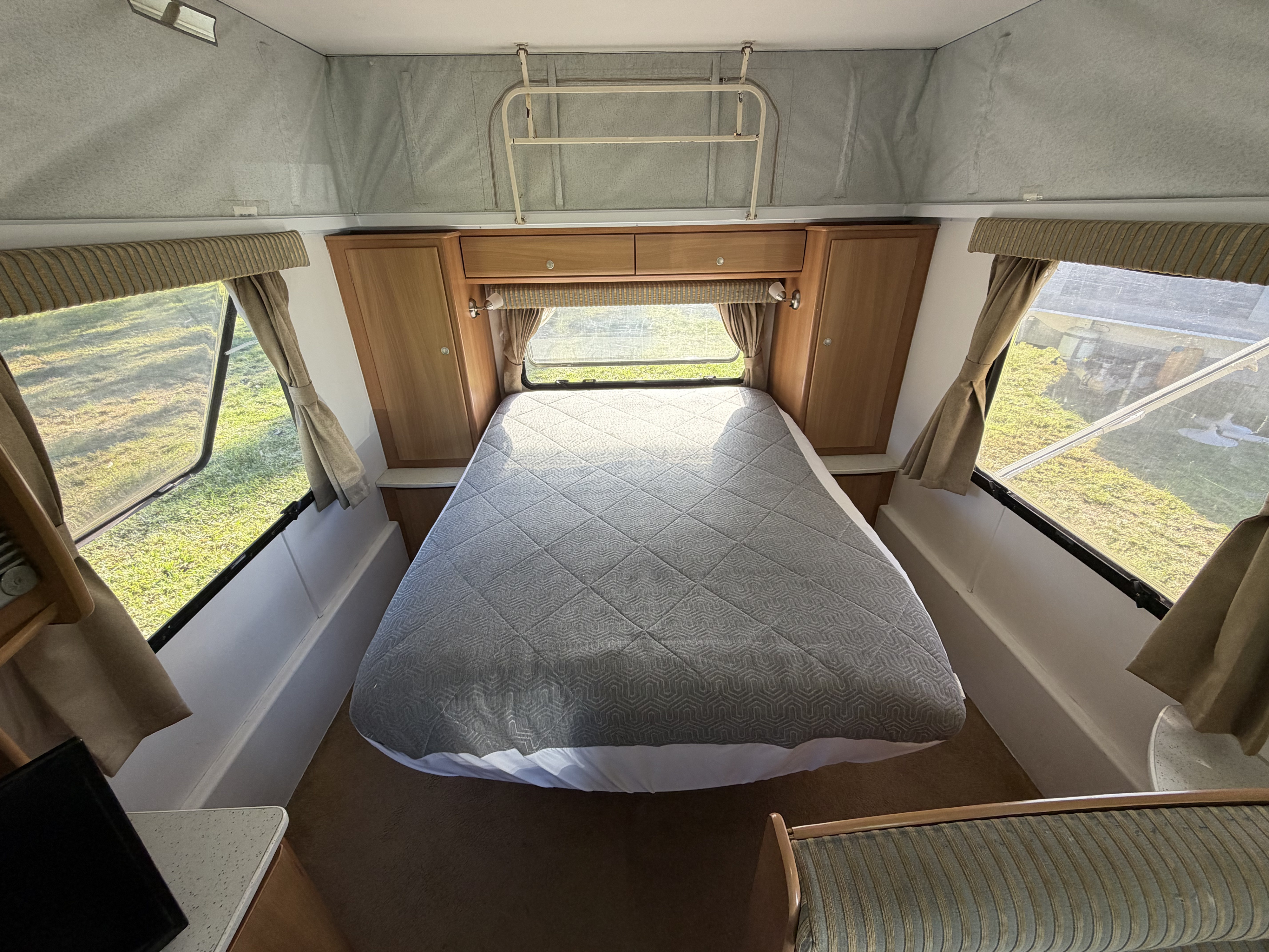 Inside an RV rental: a bright bedroom with a comfy patterned bed, surrounding windows, wooden storage, and a pop-top ceiling.. Jayco J Series 2008