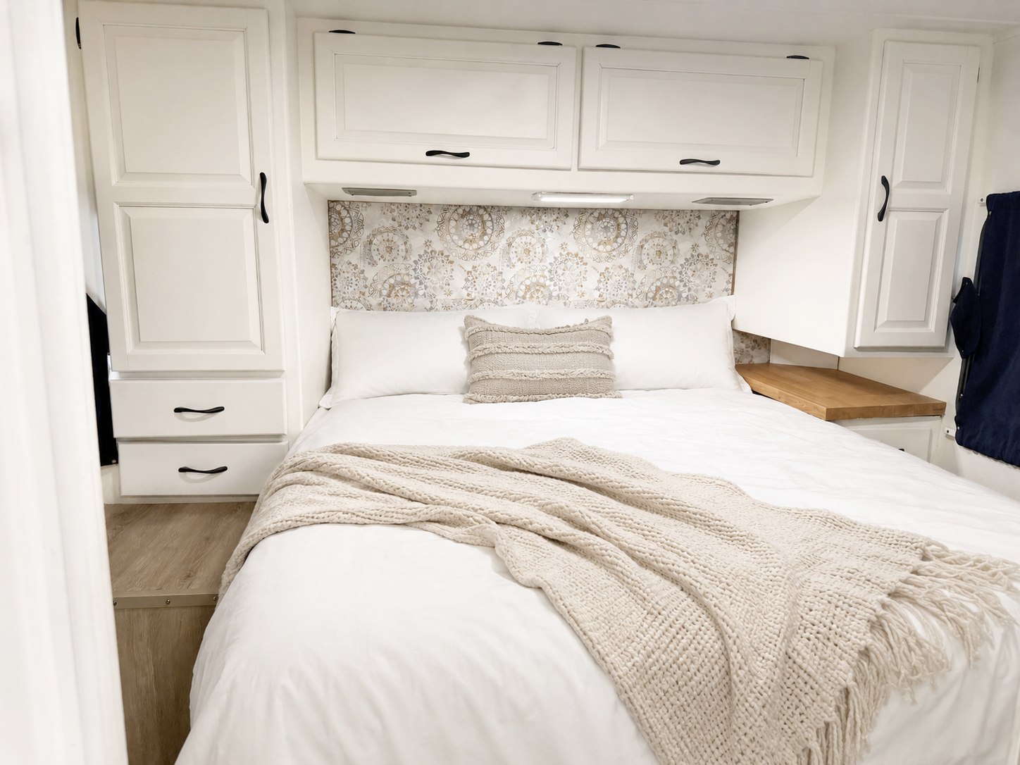 Inside a cozy RV rental bedroom: white bed with a textured throw, patterned headboard, and white storage cabinets with a wooden shelf.. Thor Majestic 2012