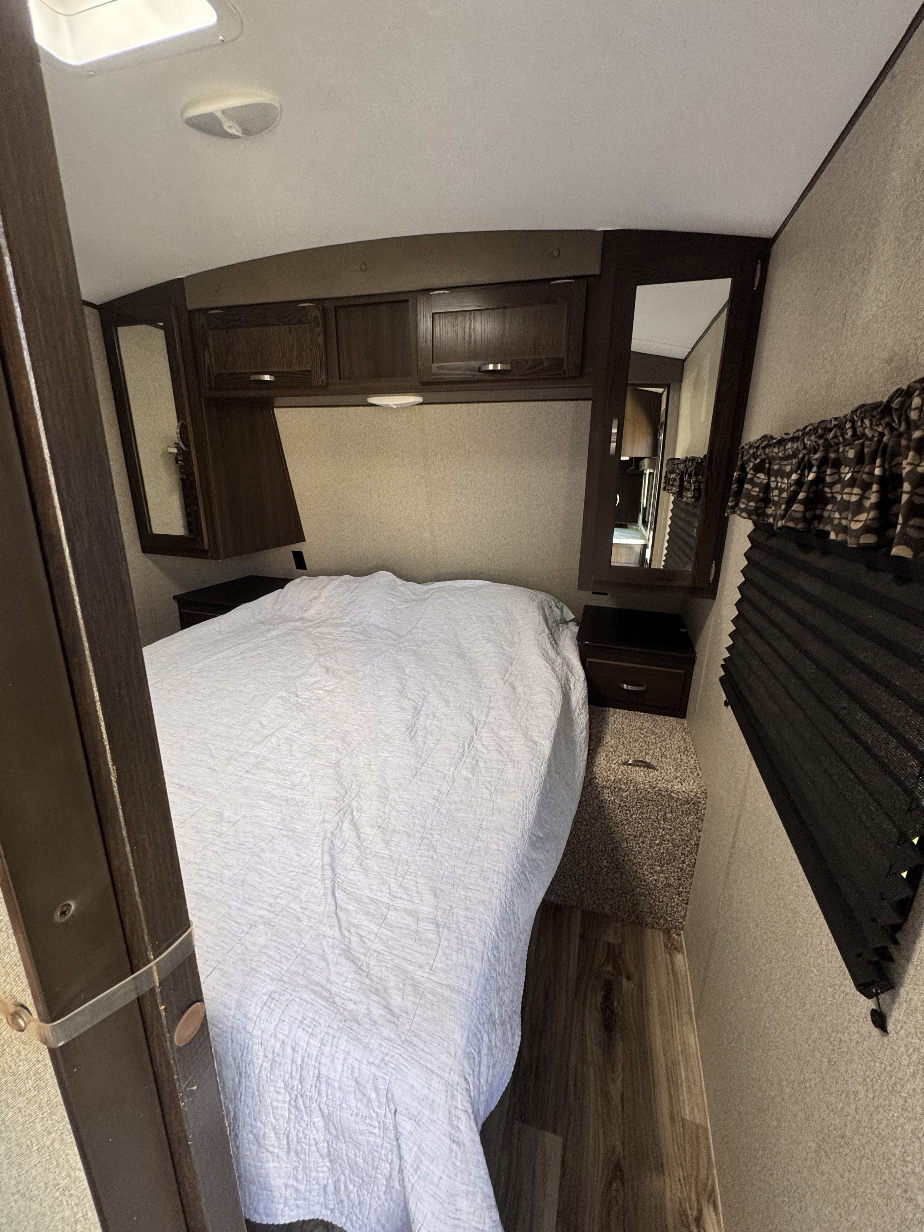 Inside an RV rental, this cozy bedroom features a white quilted bed, overhead cabinets, bedside tables, and pleated window blinds.. Keystone RV Springdale 2019