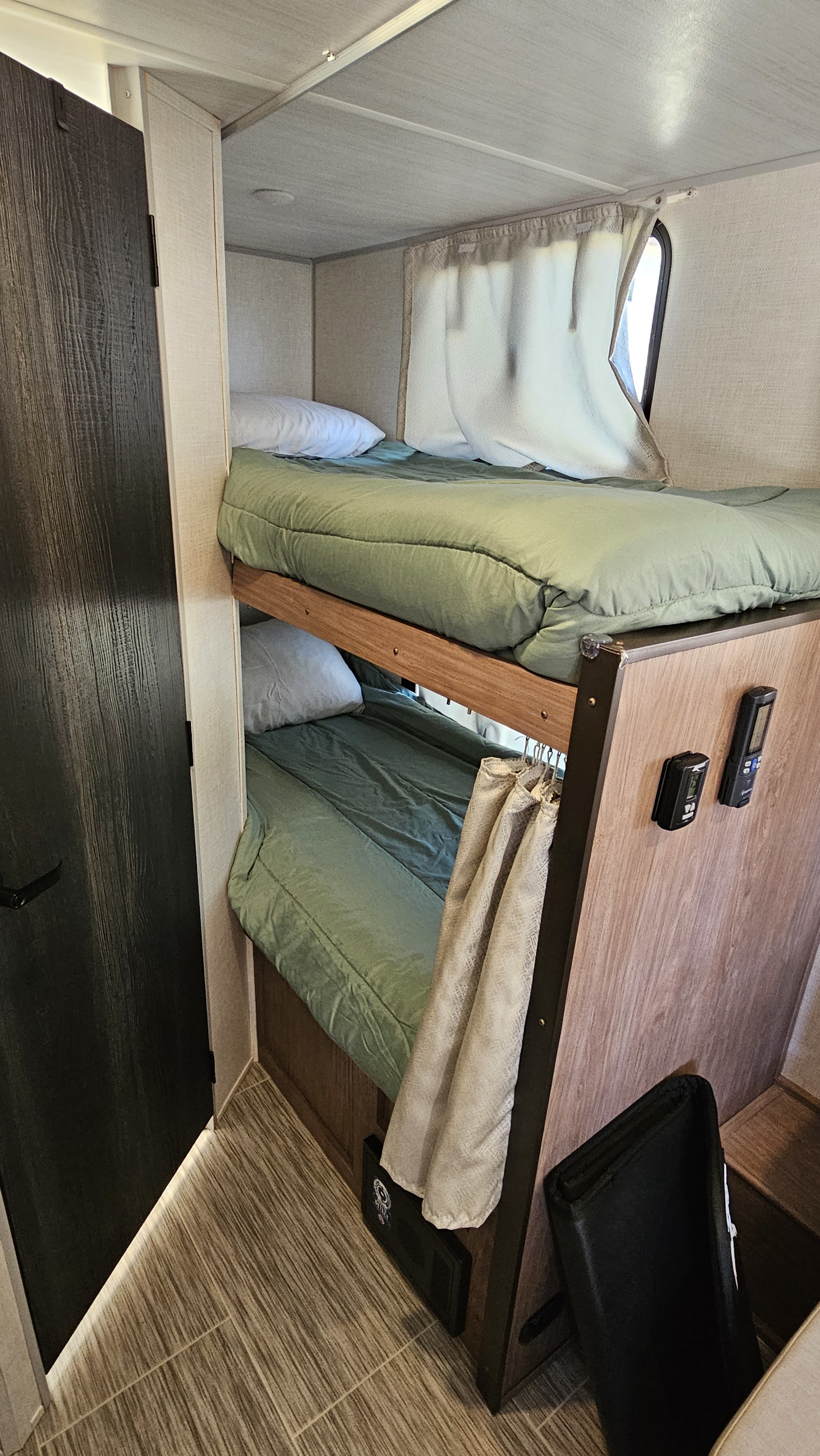 Compact rv rental interior with bunk beds, green bedding, white pillows, and a window. Door on left, controls on side.. No Boundaries 19 2022