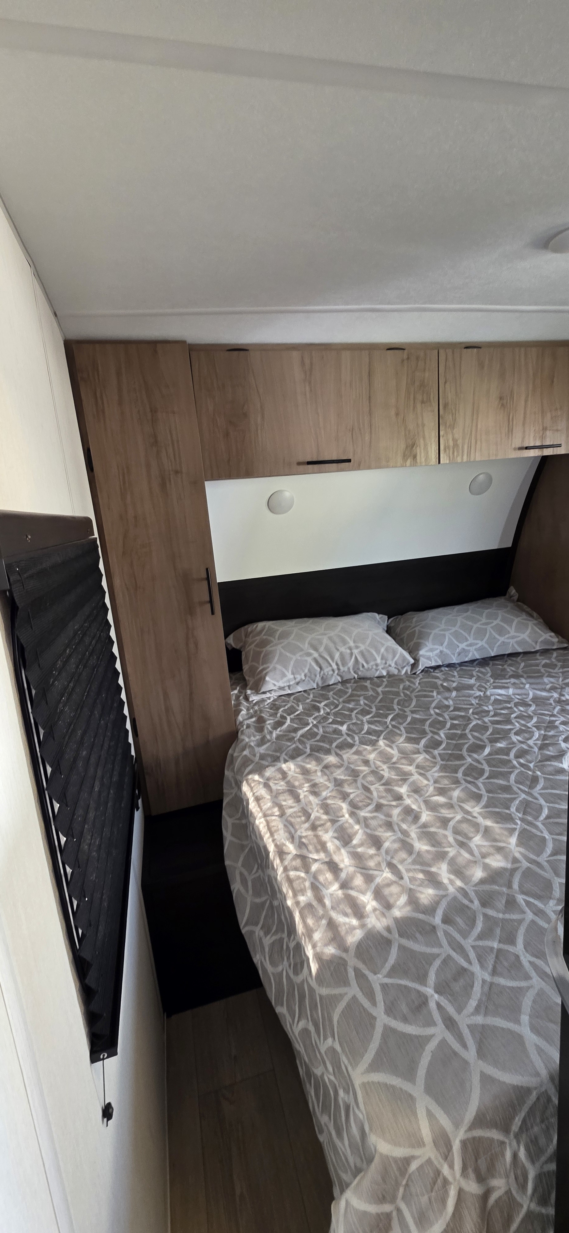 Compact RV rental bedroom featuring a patterned bed, overhead wood cabinets, and a pleated window blind, ready for adventure.. Forest River Cherokee Grey Wolf 2025