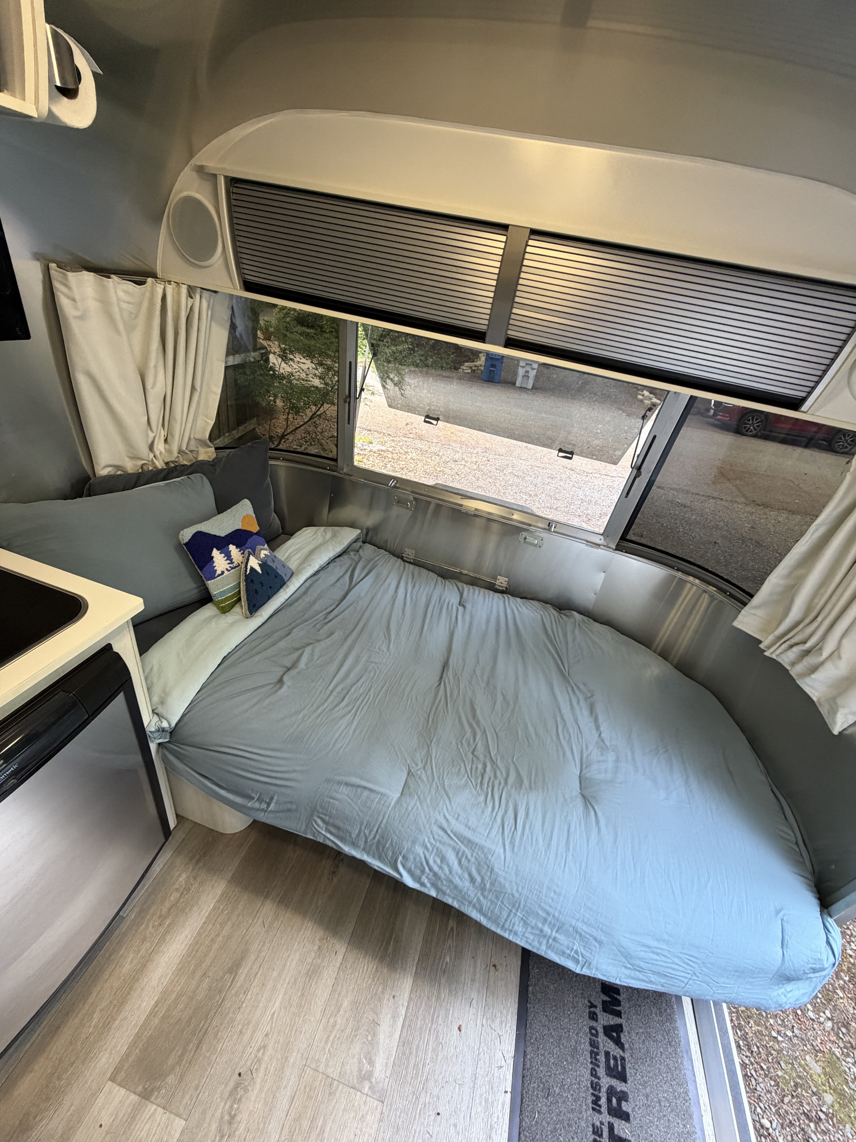View inside a modern Airstream RV rental: a bed with blue linens, large windows, and a compact kitchen area.. Airstream Sport 2016