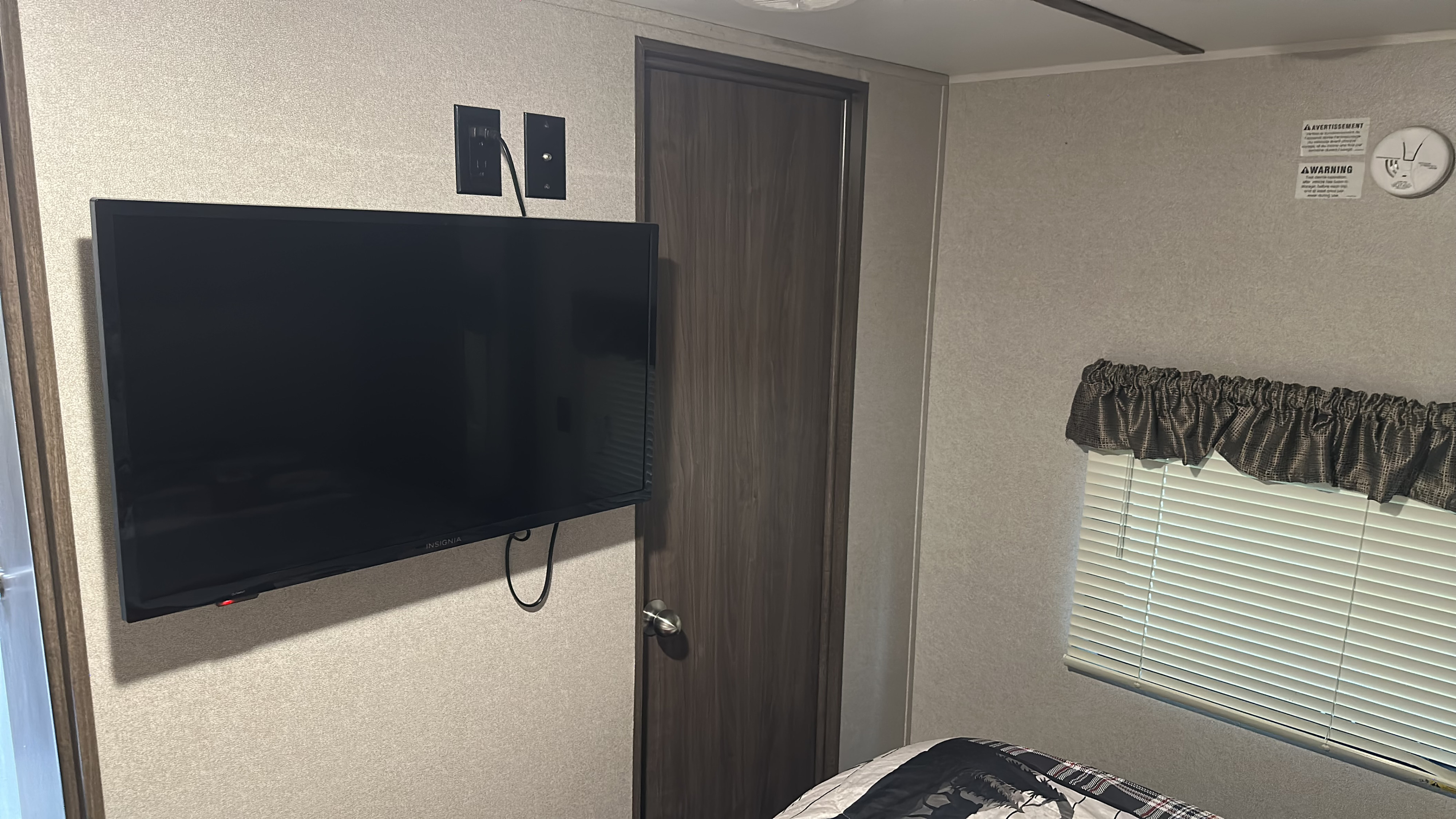 An RV rental bedroom with a wall-mounted TV, power/cable outlets, door, window with blinds, and bed. Smoke detector.. Keystone RV Hideout Luxury 2019
