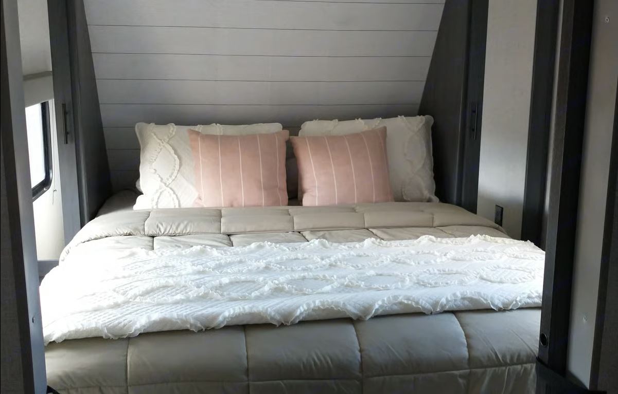 Cozy RV rental bedroom: a made bed with soft white and pink pillows, white ruffled blanket, and a window to the left.. Aspen Trail LE 2021