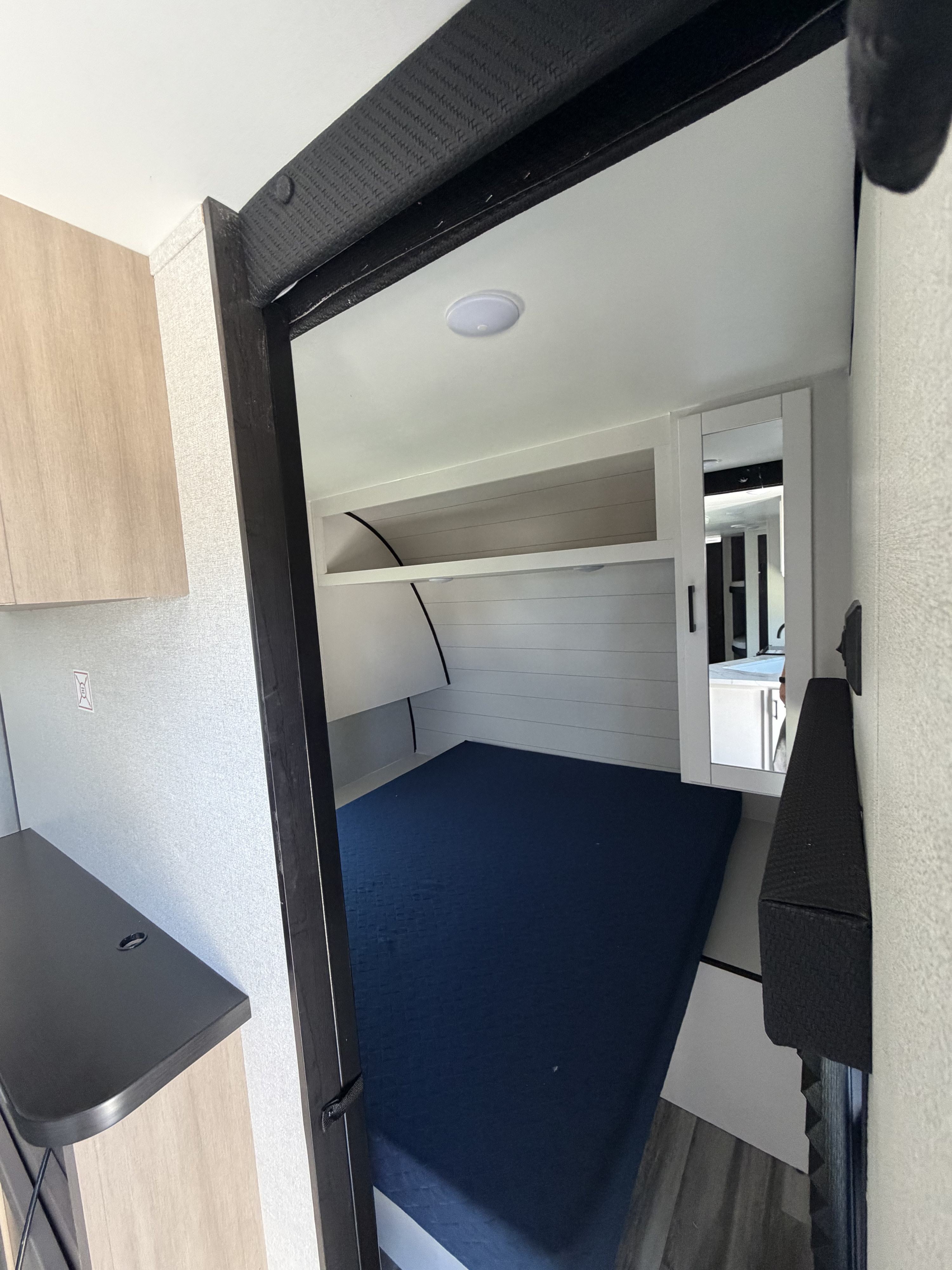 Compact RV rental bedroom featuring a blue bed, white shiplap walls, overhead storage shelf, and a mirrored cabinet.. Jayco Jay Flight 2027