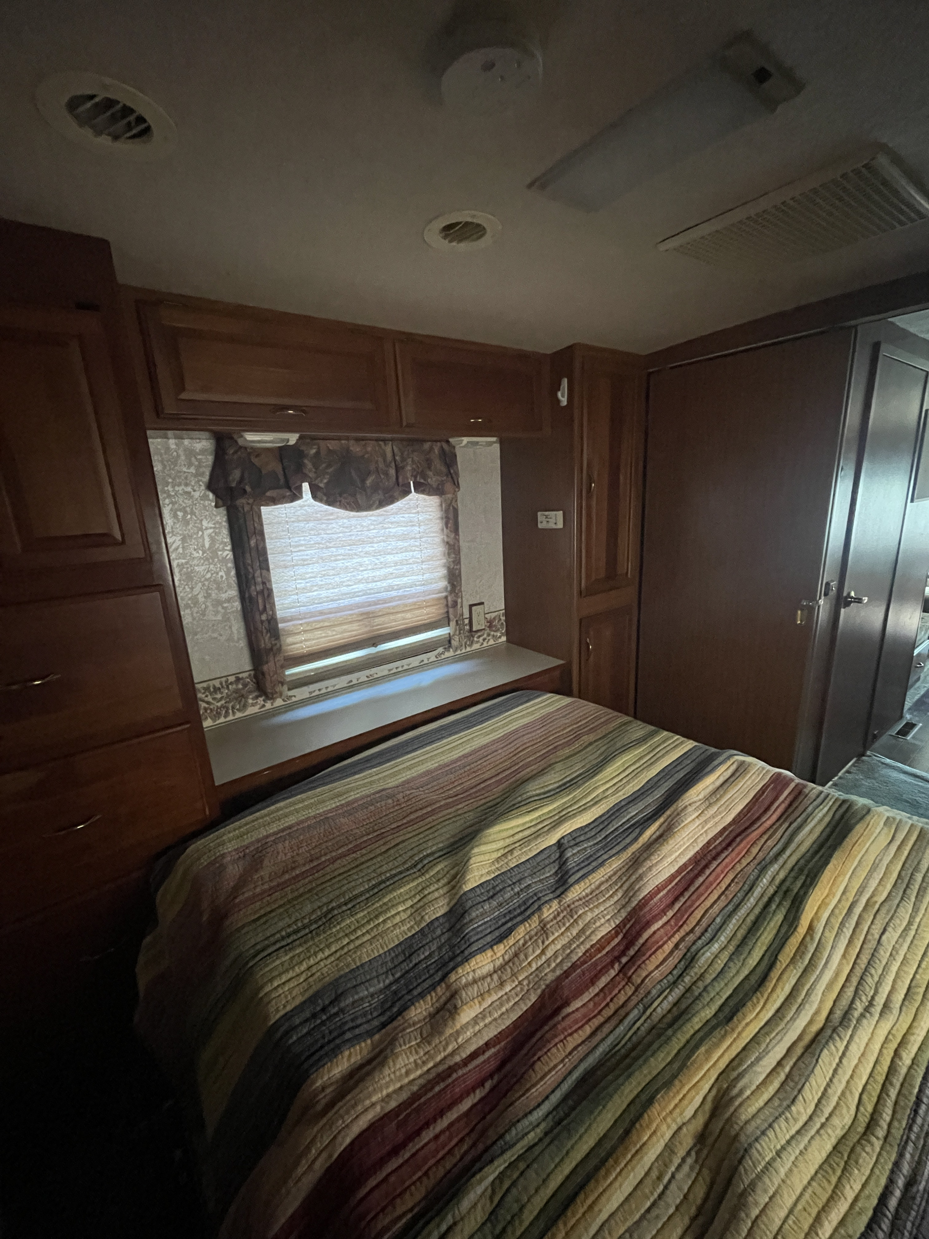 This RV rental bedroom features a striped bed, wood cabinetry, a window with blinds, and ceiling fixtures.. Winnebago Chieftain 1999