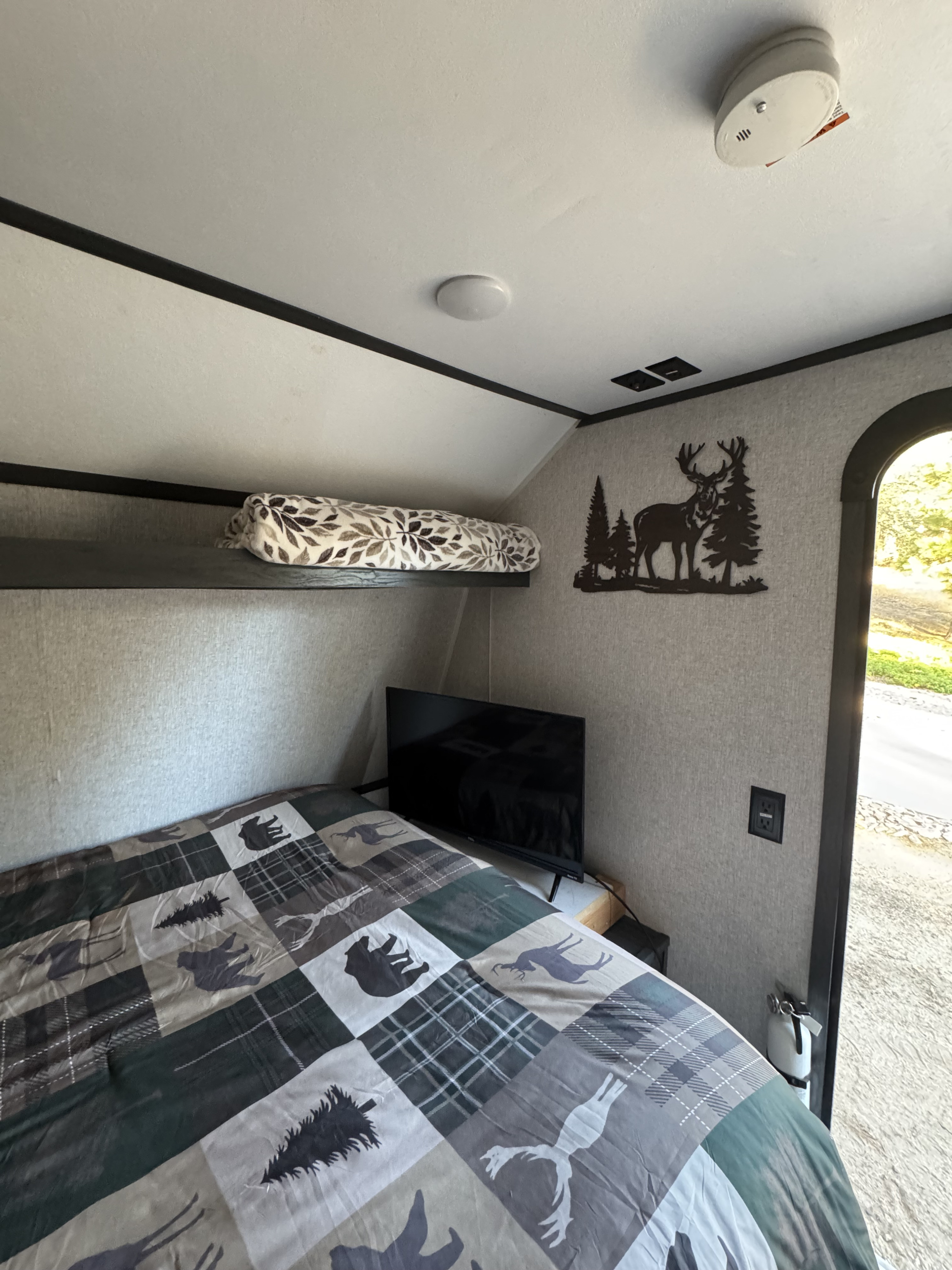 Cozy rv rental bedroom featuring forest-themed bedding, a small TV, and rustic deer wall art. Safety items like a smoke detector are visible.. Aspen Trail Aspen Trail Trailer 2022
