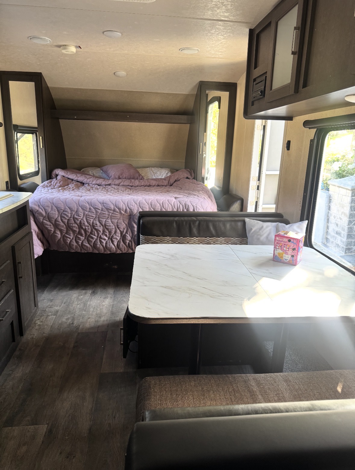 This rv rental interior features a cozy purple bed, a modern dining area with a marble-patterned table, and ample windows.. Forest River Wildwood X-Lite 2019