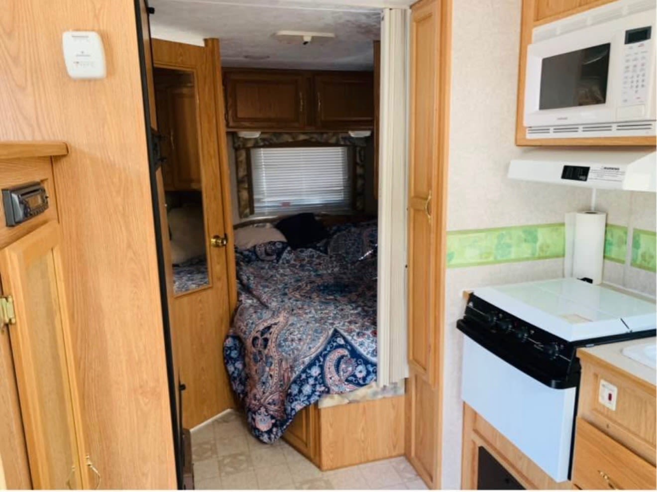 View inside an RV rental showing a bedroom with a patterned bed, window, and wood cabinets, next to a kitchen with a microwave and stove.. Komfort Trailblazer 2003