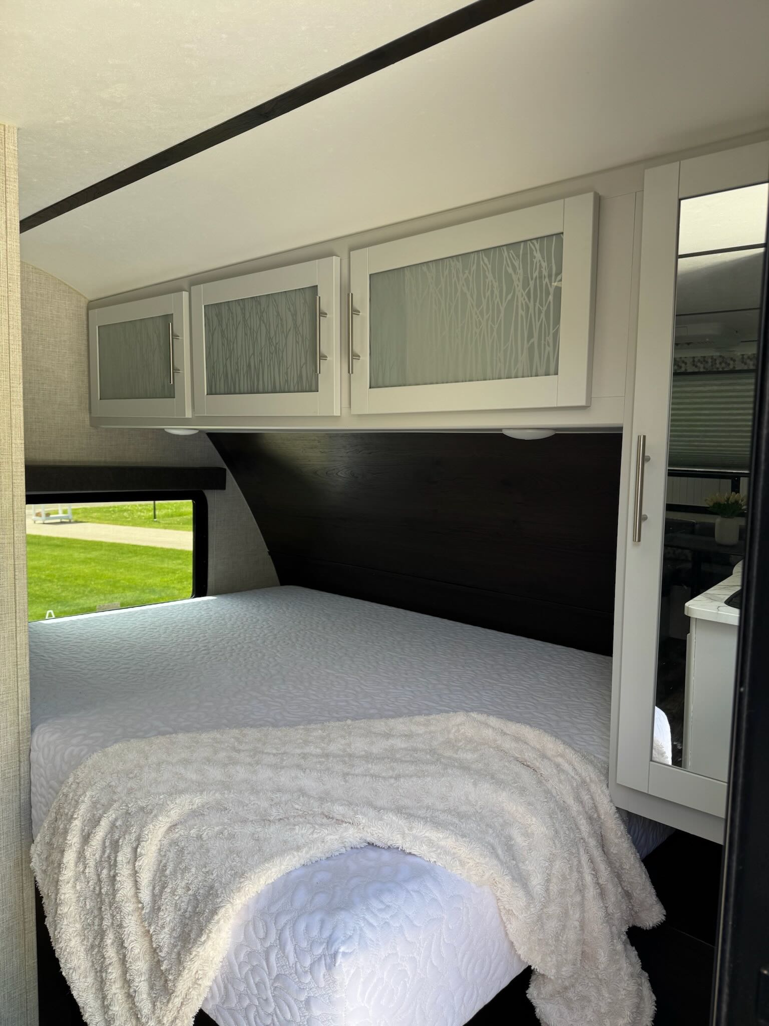 Cozy RV rental bedroom featuring a bed with white bedding and fluffy throw, overhead cabinets, and a window to nature.. Keystone RV Bullet Crossfire 2019